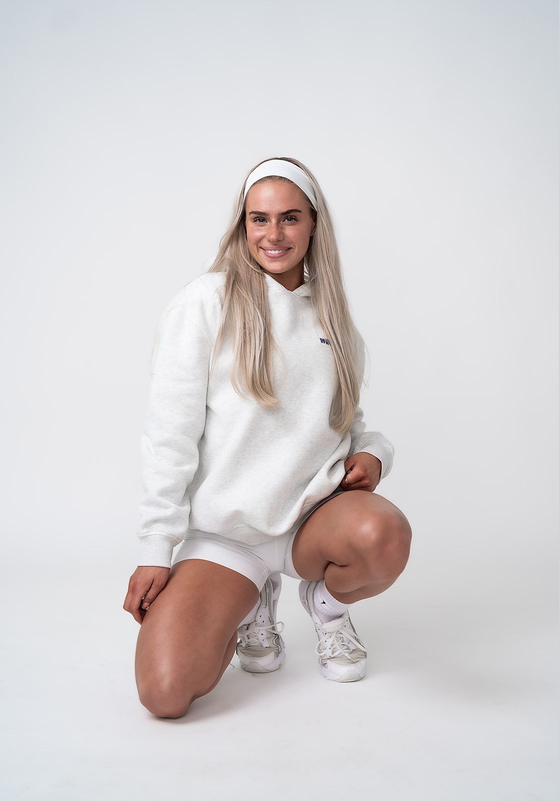 A woman in a white sweatshirt and shorts is squatting down on a white background.