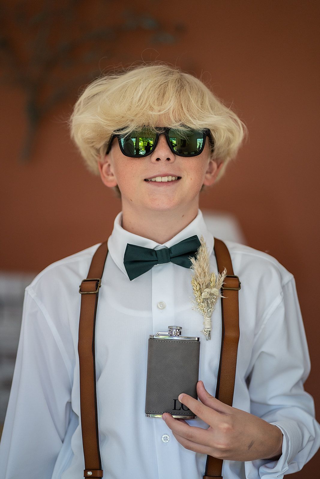 A young boy wearing suspenders and sunglasses is holding a flask.
