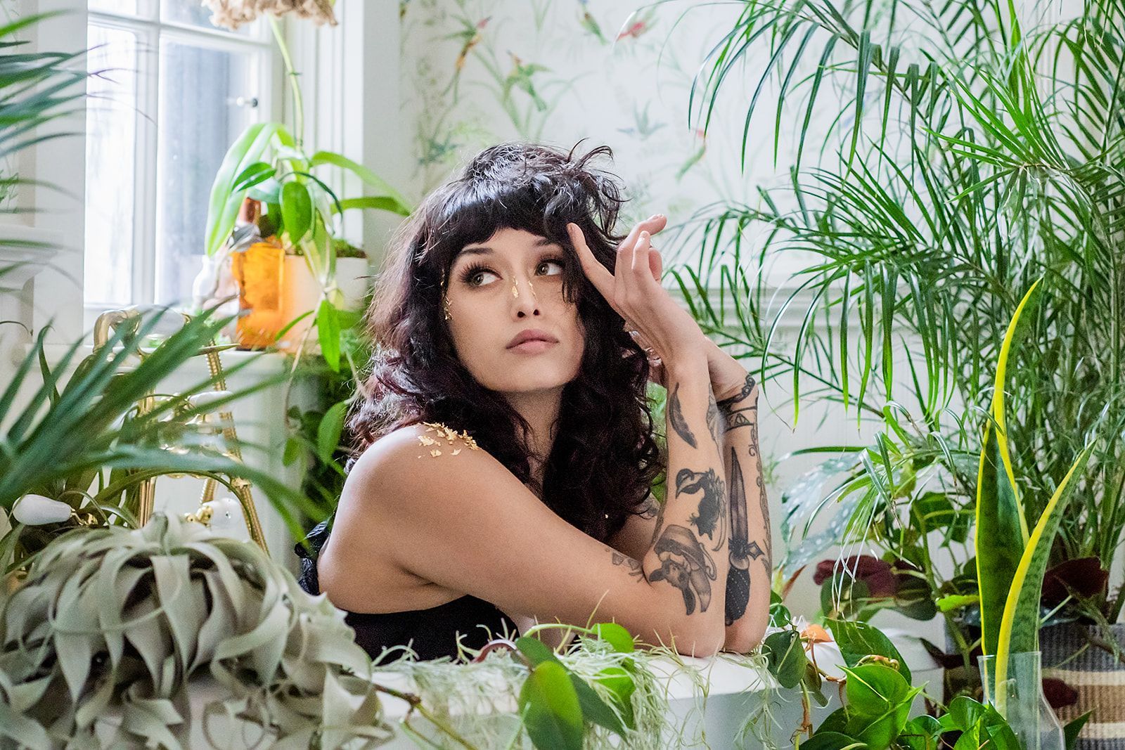 A woman with tattoos is sitting in a room surrounded by plants.
