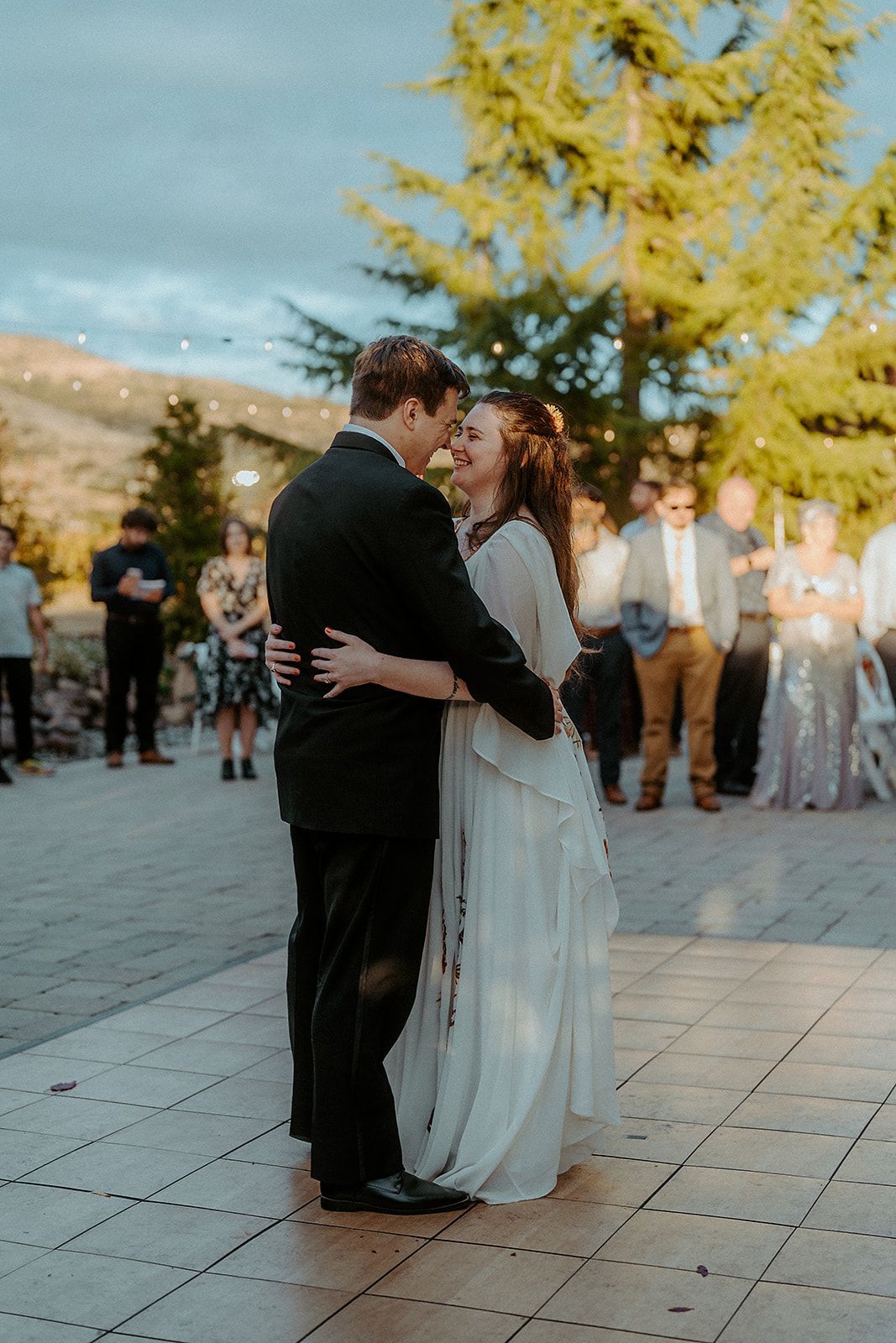 Couple dances at wedding reception; outdoors, smiling, evening light, guests in background.