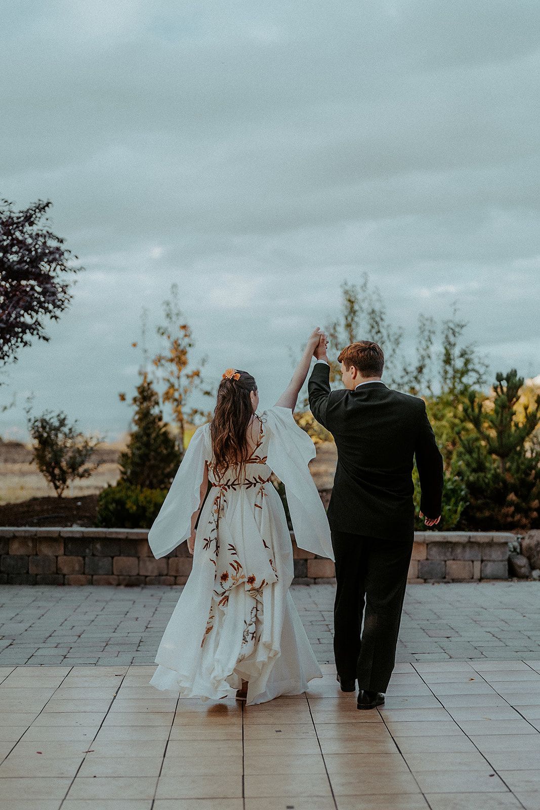 Bride and groom walk hand-in-hand, backs to the camera, on a patio. Cloudy sky. Bride's dress has floral accents, groom in a suit.