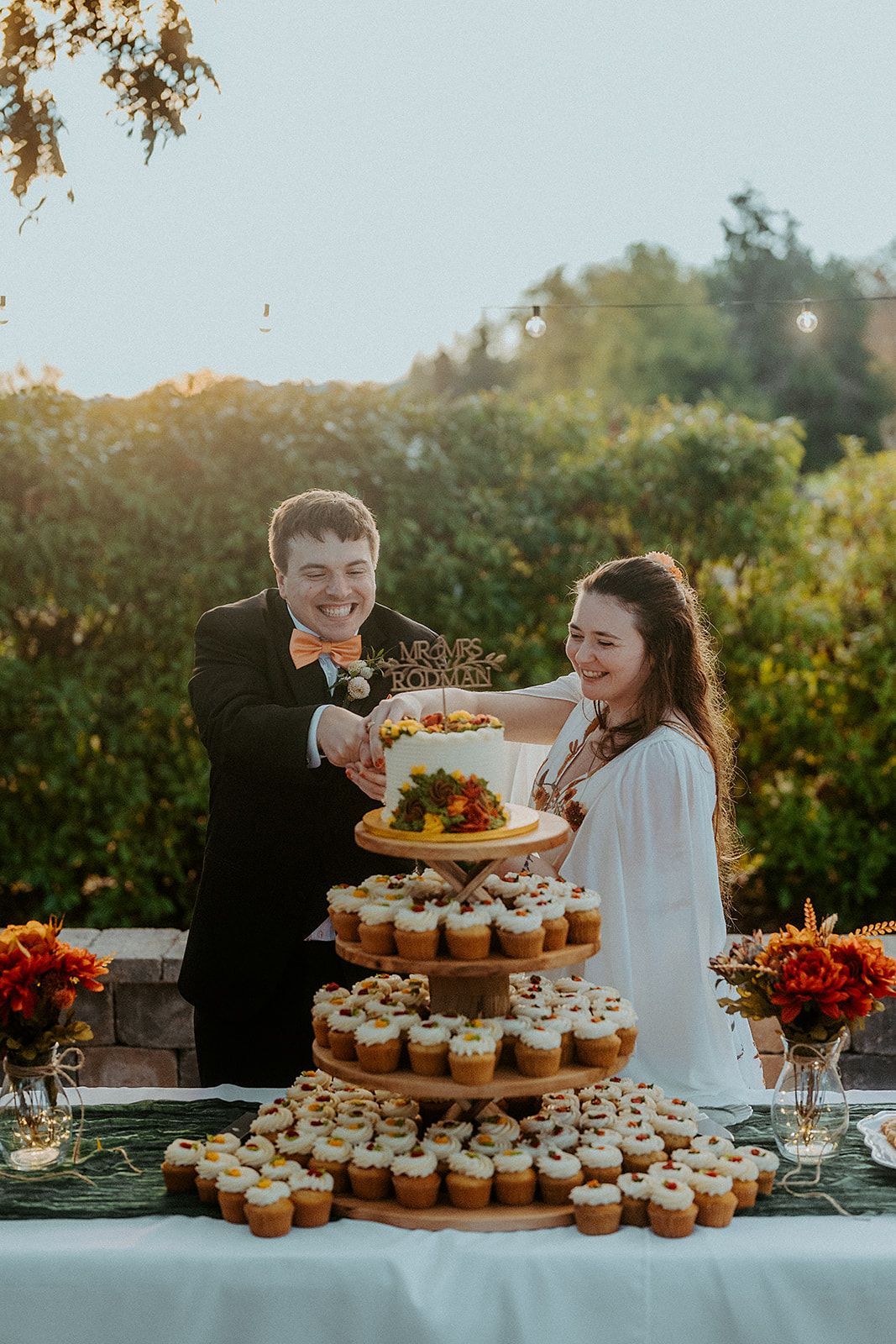A bride and groom are cutting their wedding cake in front of a table filled with cupcakes.