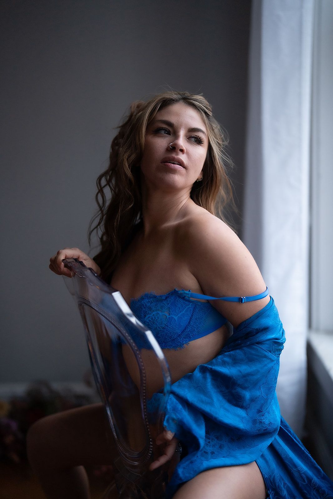 A woman in blue lingerie is sitting on a clear chair in front of a window.