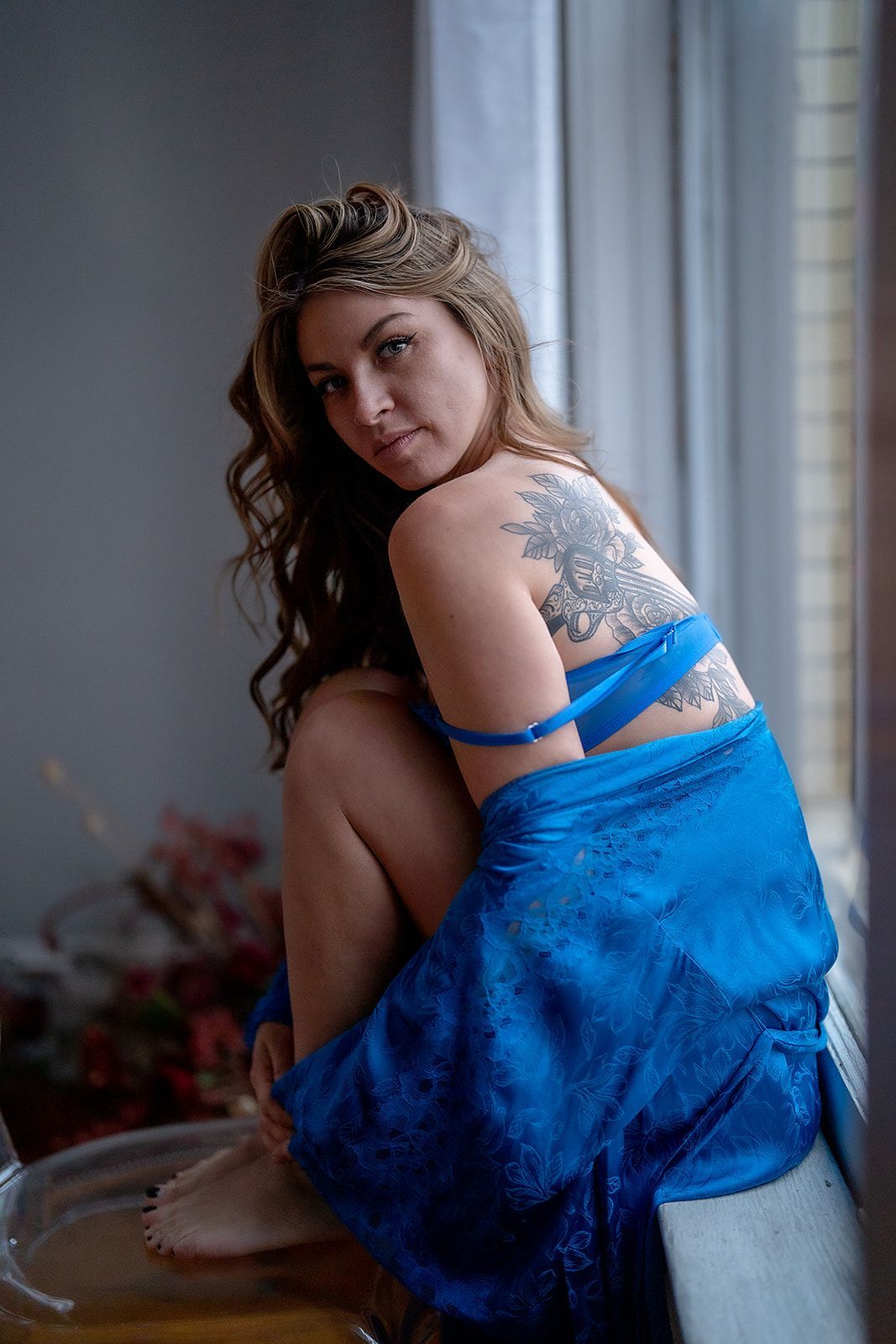 A woman in blue lingerie is sitting on a chair in front of a window.