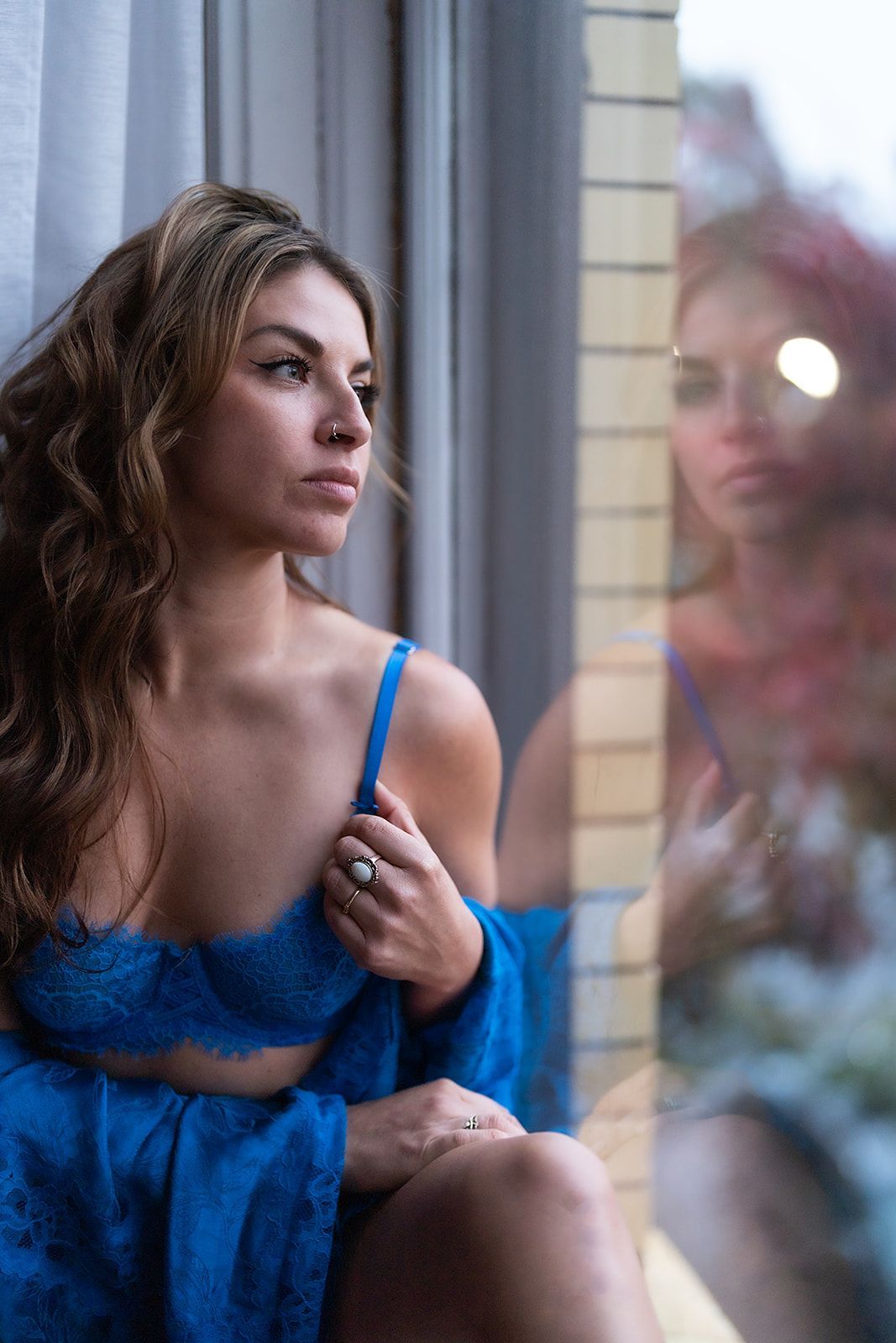 A woman in lingerie is sitting on a window sill looking out the window.