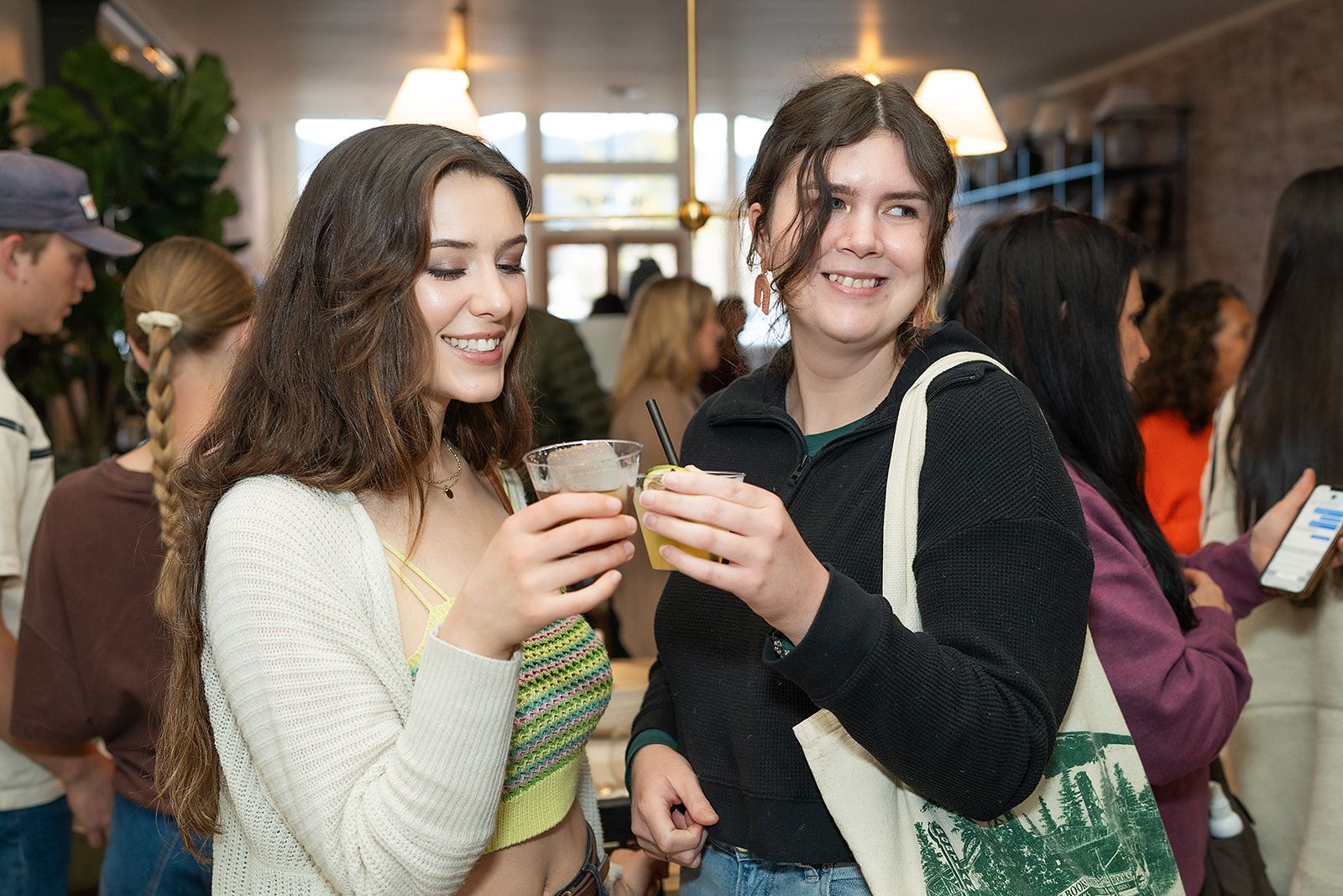 Two women are toasting with drinks in a crowded room.