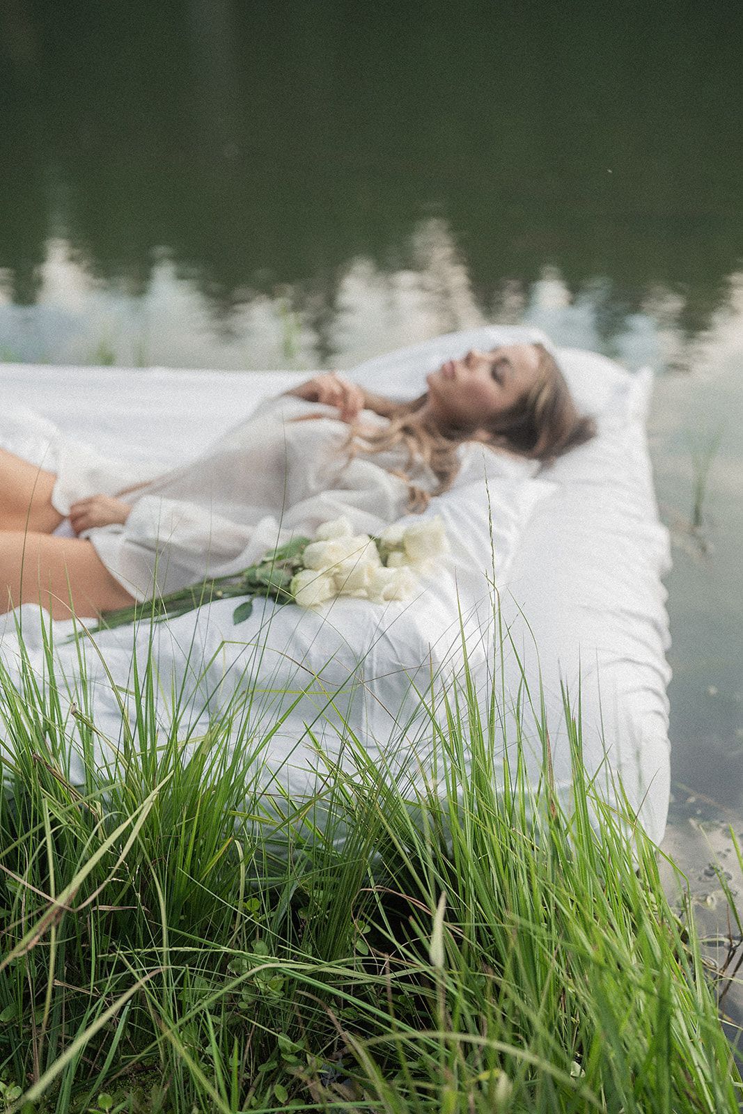 A woman is laying on a bed in the grass next to a body of water.