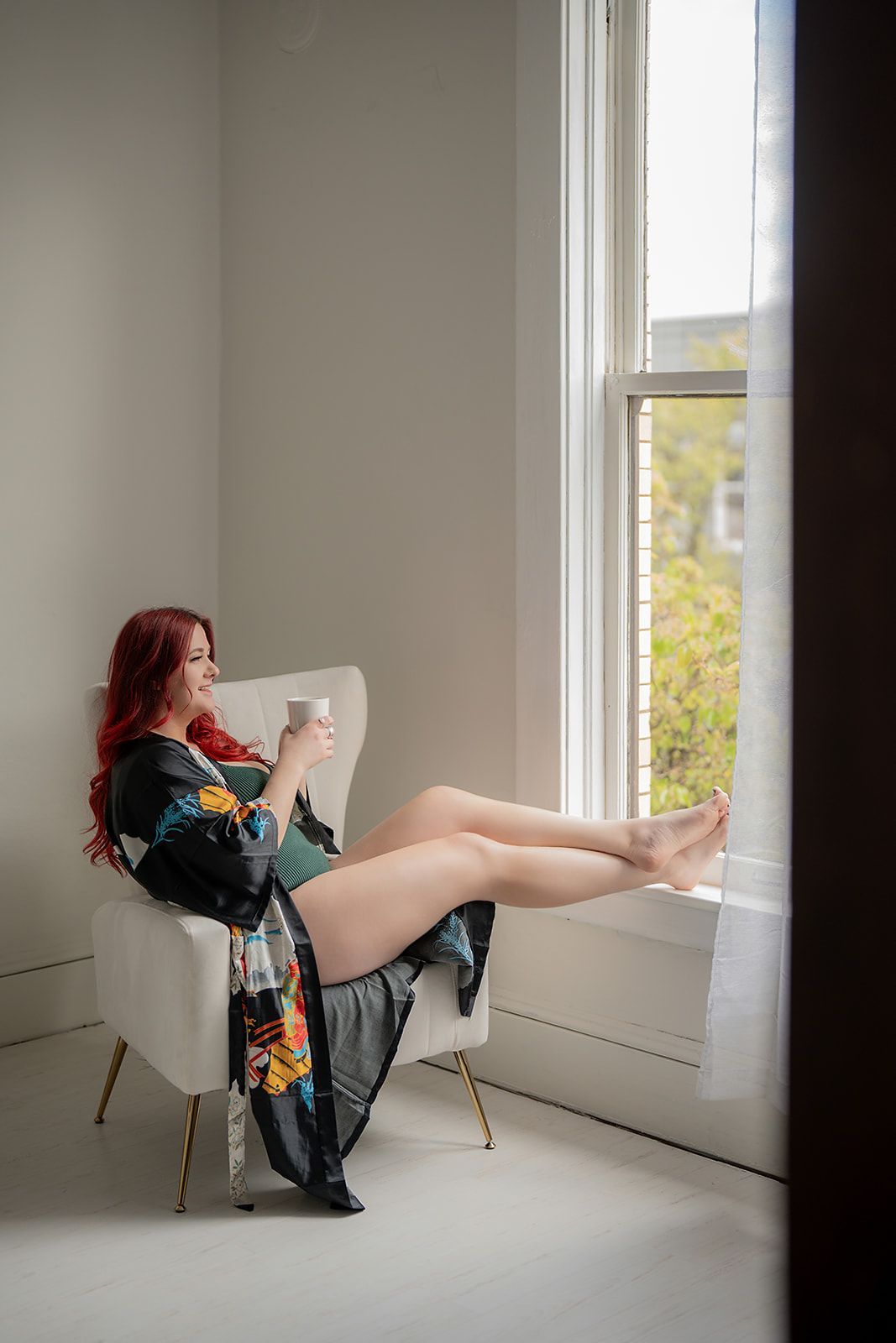 A woman is sitting in a chair in front of a window holding a cup of coffee for a boudoir photo shoot in Medford, Oregon