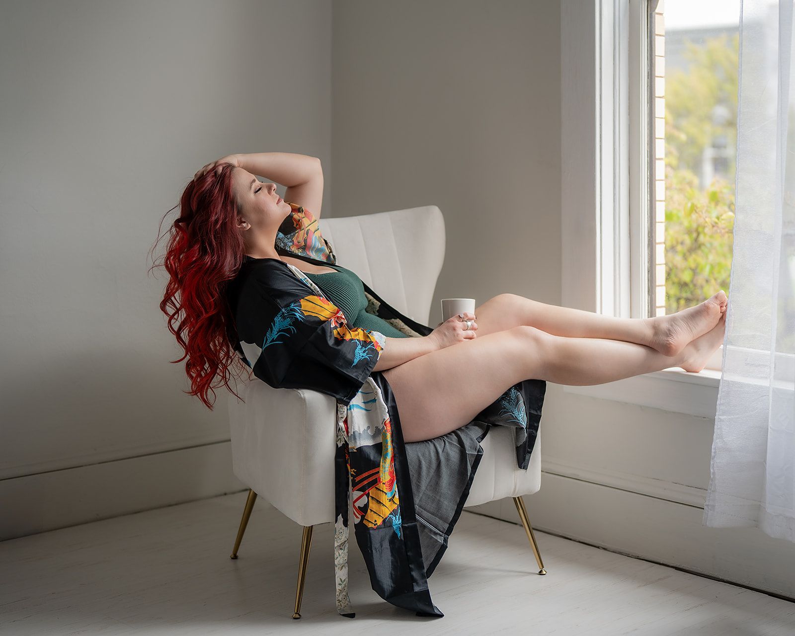A woman is sitting in a chair looking out a window for a boudoir photo shoot in Medford, Oregon