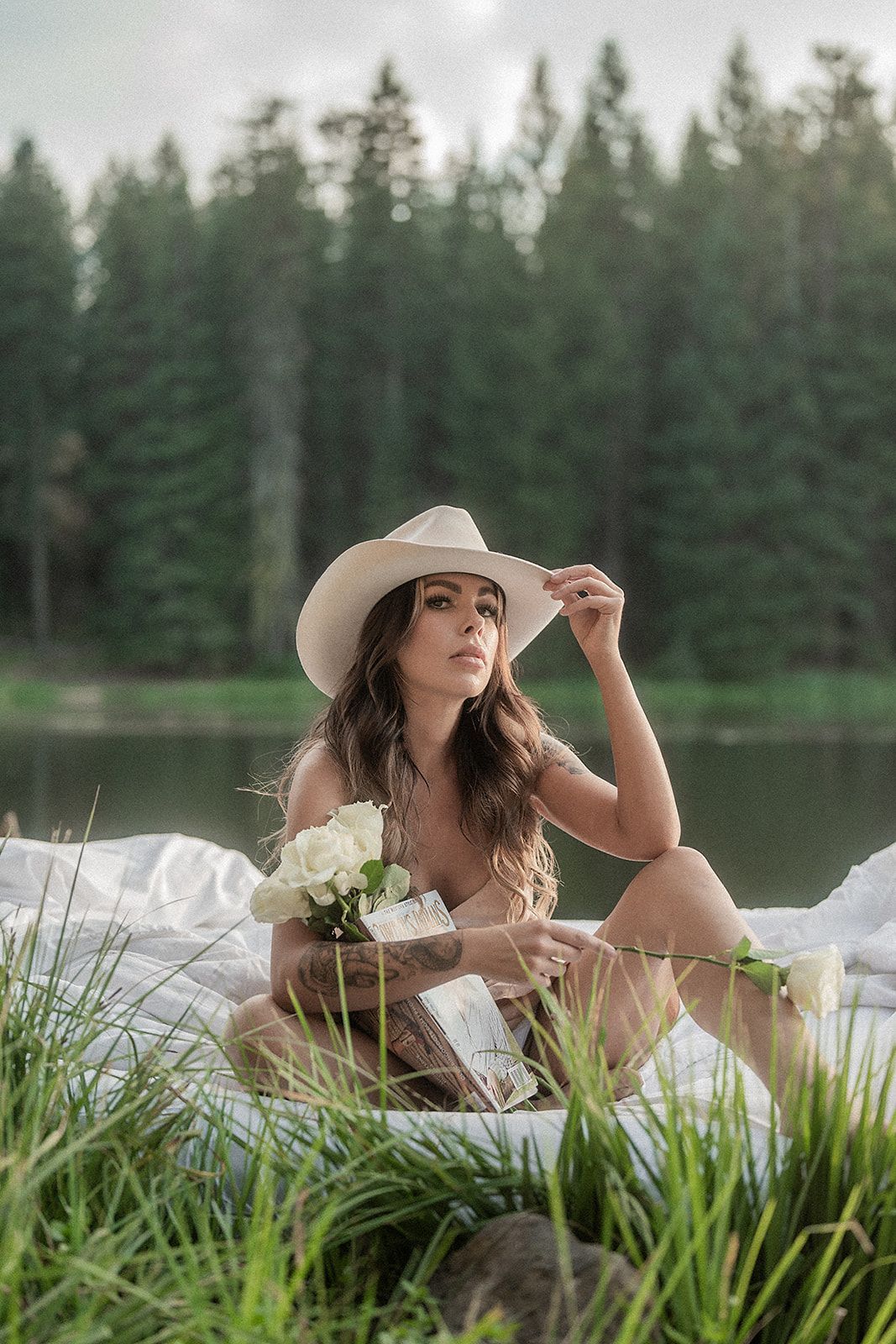 A woman in a cowboy hat is sitting on a bed next to a lake.