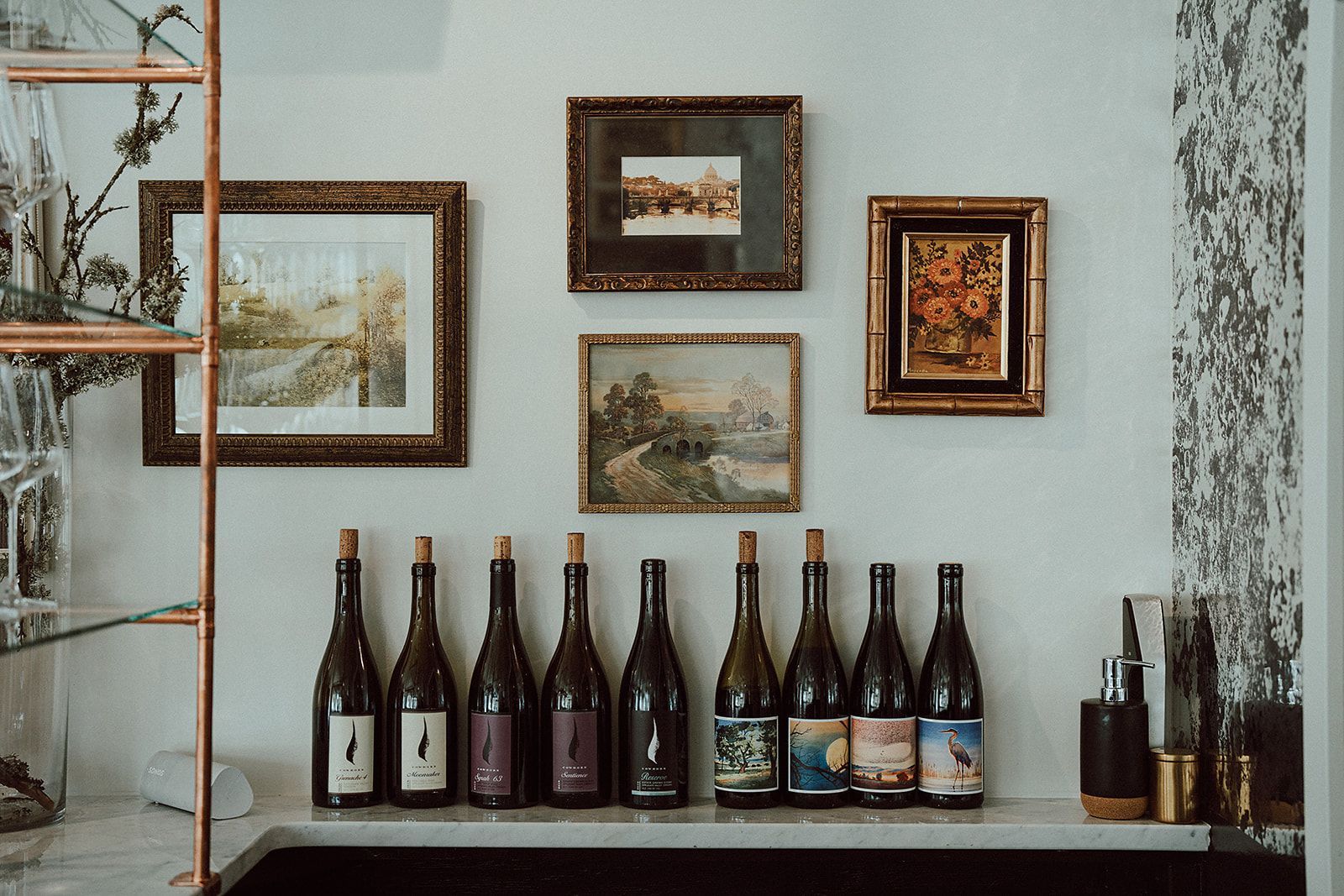 A row of wine bottles sitting on top of a counter next to a wall with pictures on it.