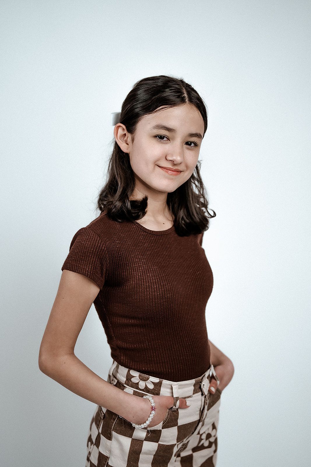 A young girl in a brown shirt and checkered skirt is standing with her hands in her pockets.