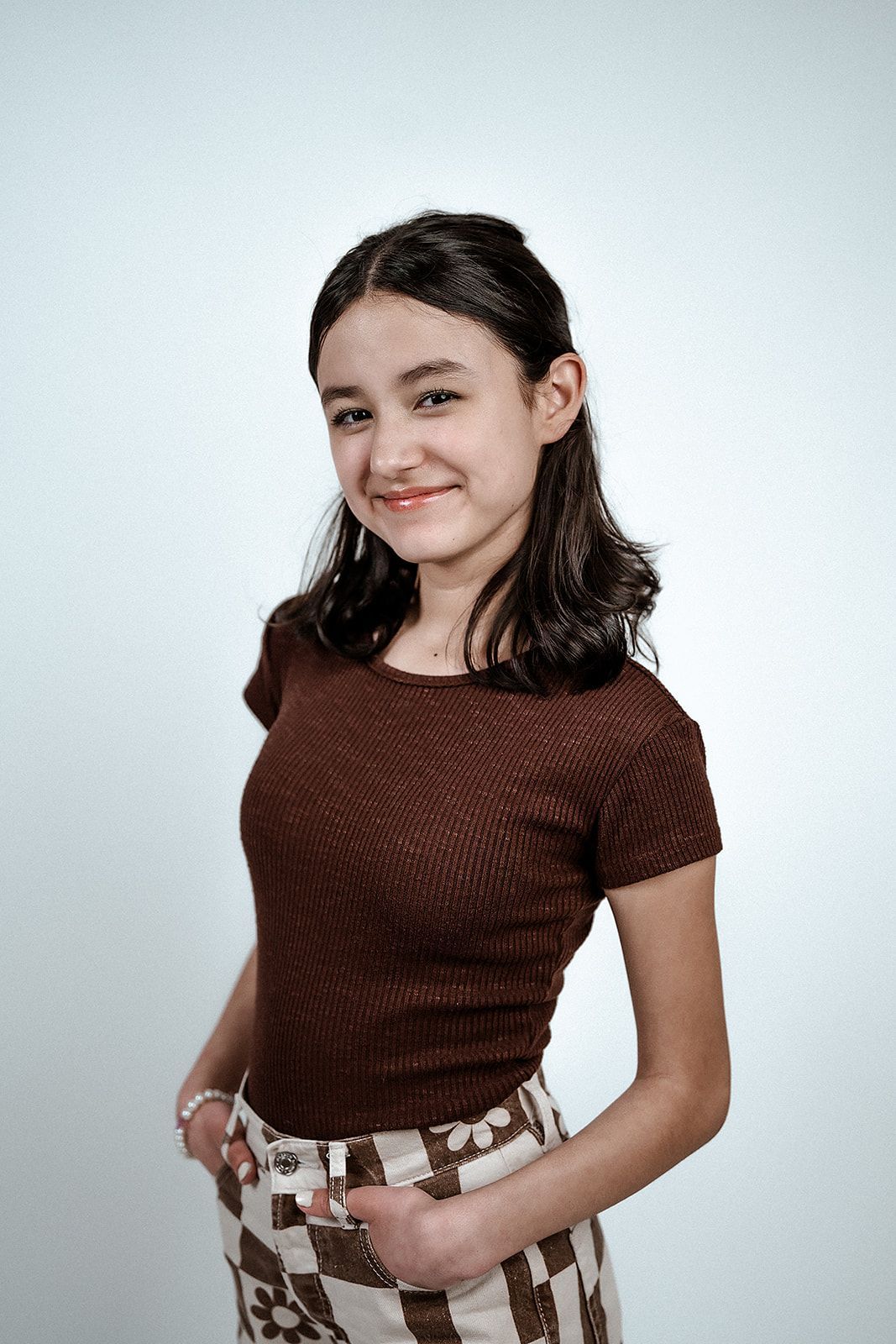A young woman in a brown top and checkered skirt is standing in front of a white wall.
