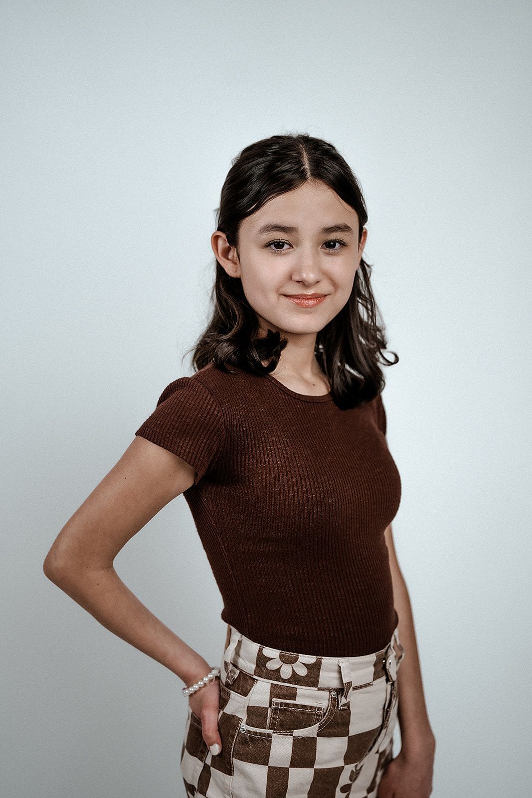 A young woman in a brown top and checkered skirt is standing in front of a white wall.
