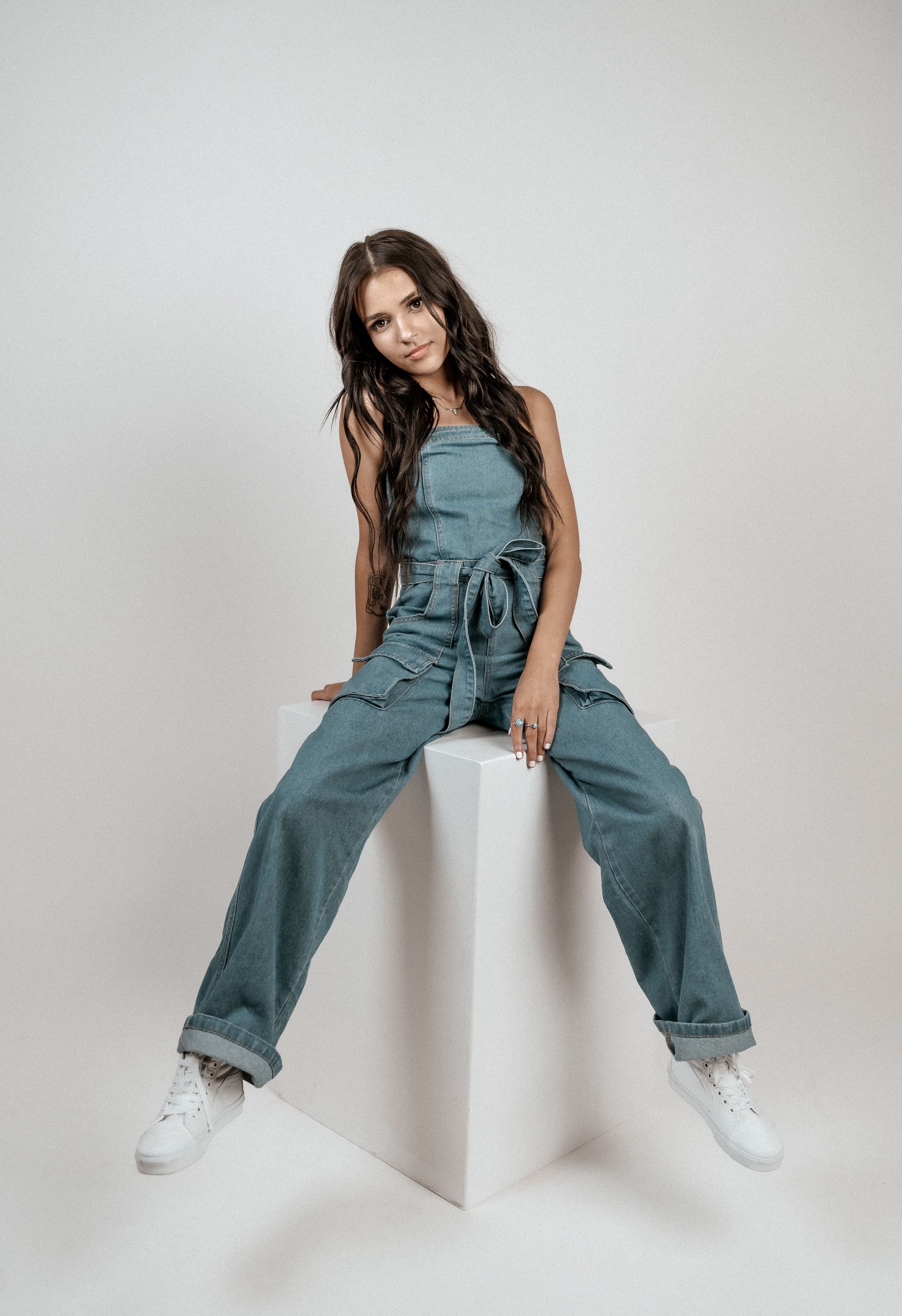 A woman in overalls is sitting on a white cube.