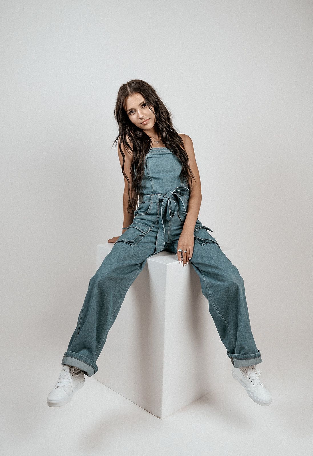 A woman in a denim jumpsuit is sitting on a white cube.