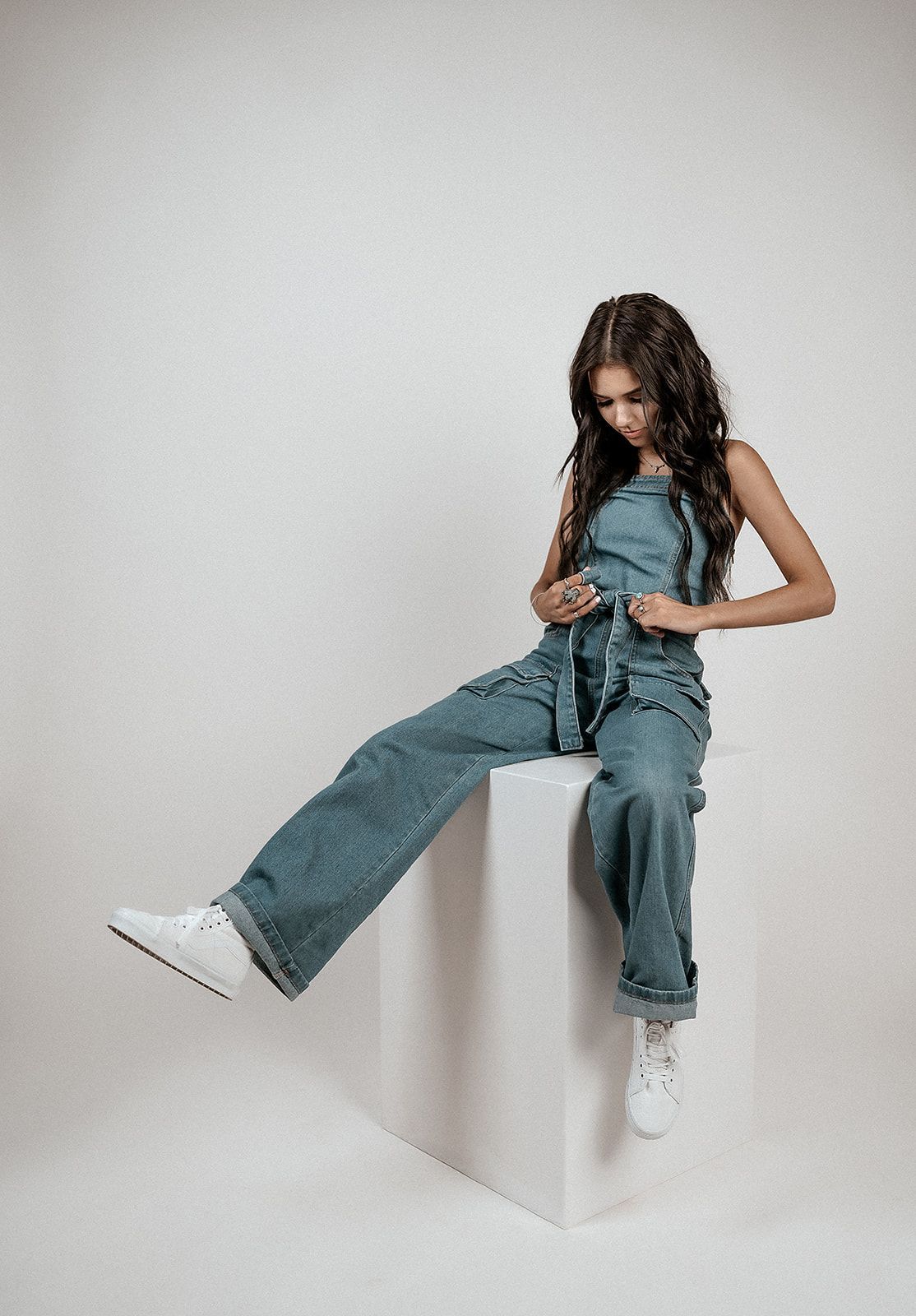 A woman in overalls is sitting on a white cube.