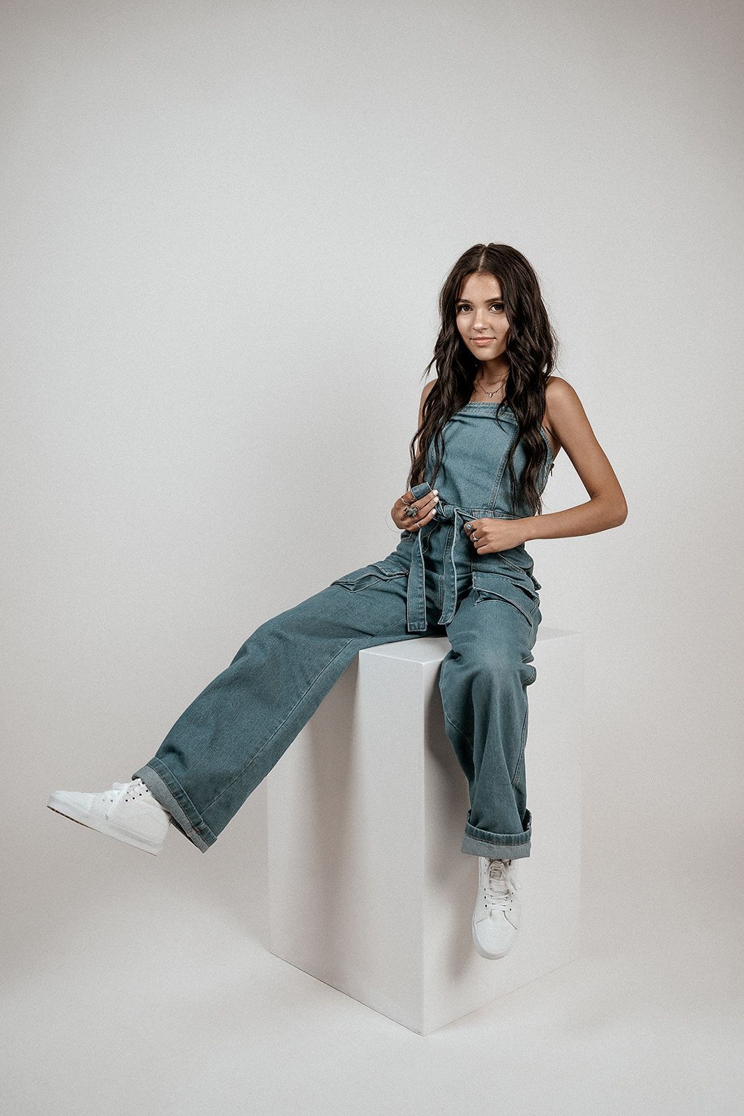 A woman in overalls is sitting on a white cube.