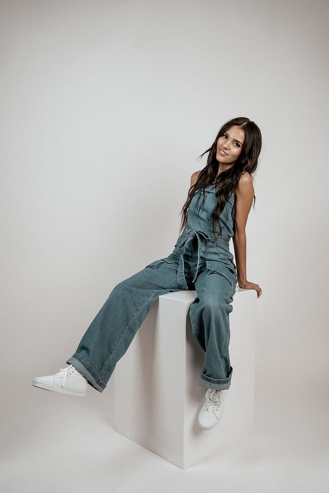 A woman in overalls is sitting on a white cube.