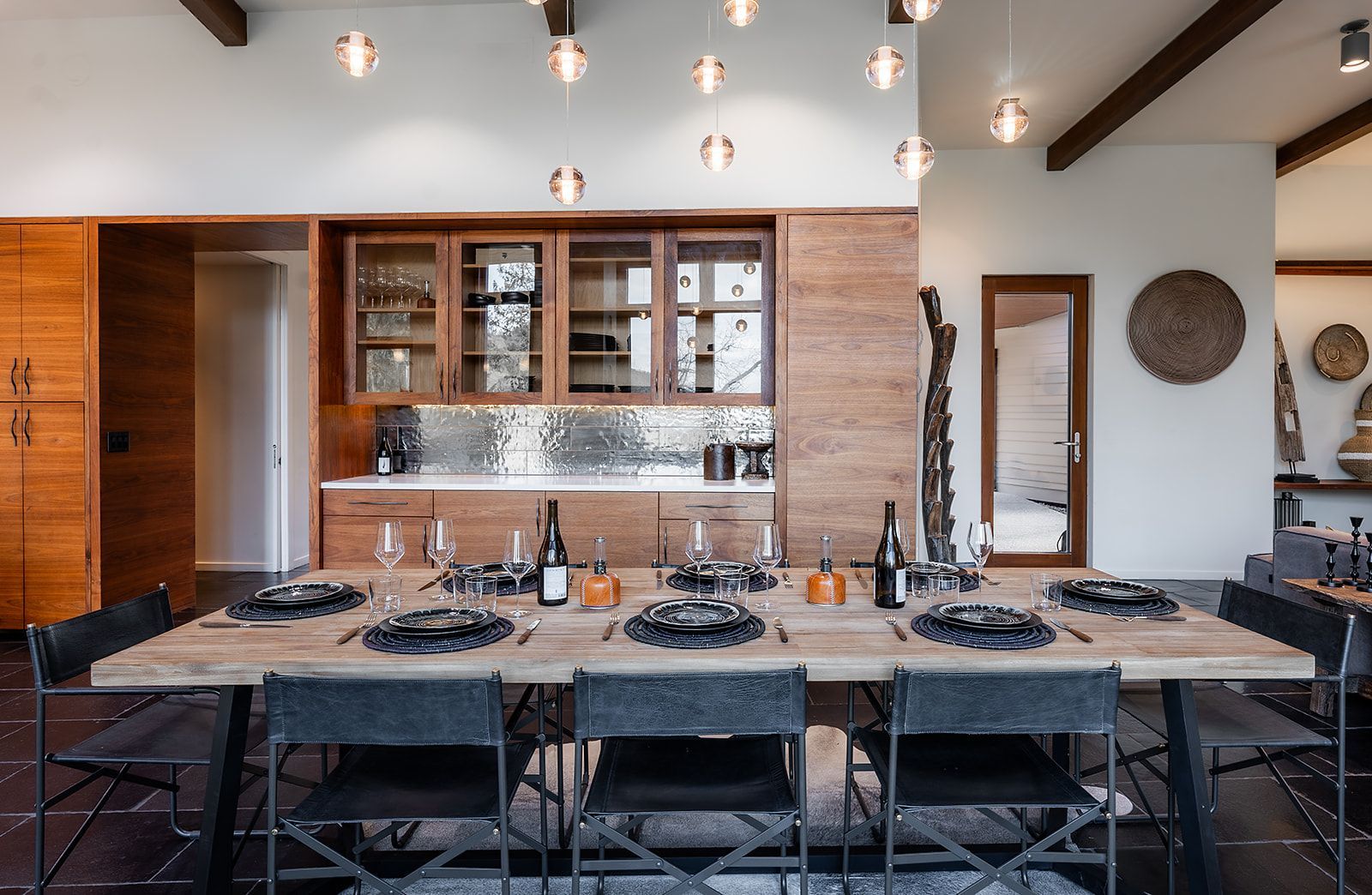 A dining room with a long table and chairs and a kitchen in the background.