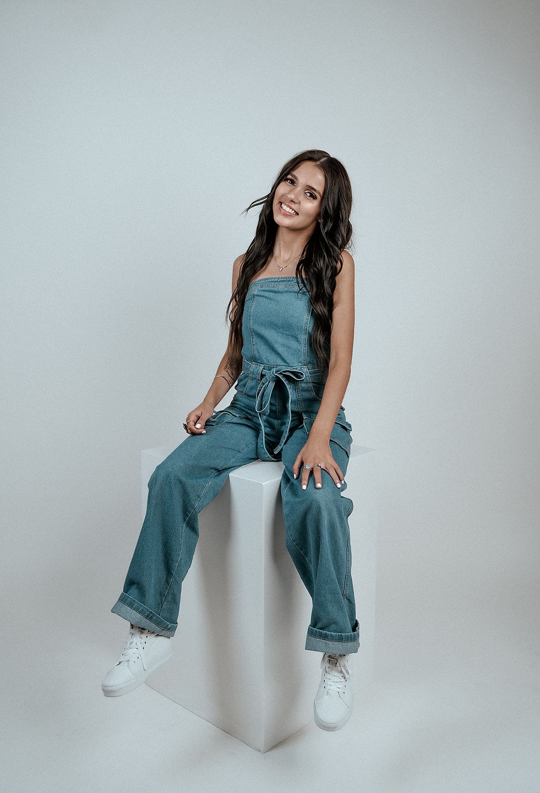 A woman in a denim jumpsuit is sitting on a white cube.