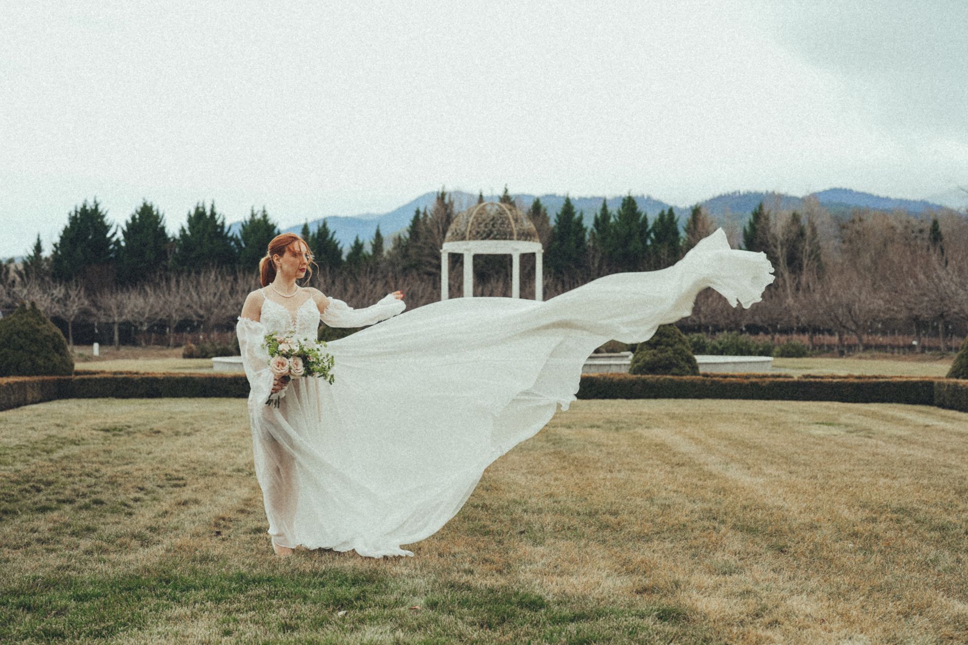 A bride in a long white dress is standing in a field with her dress blowing in the wind.