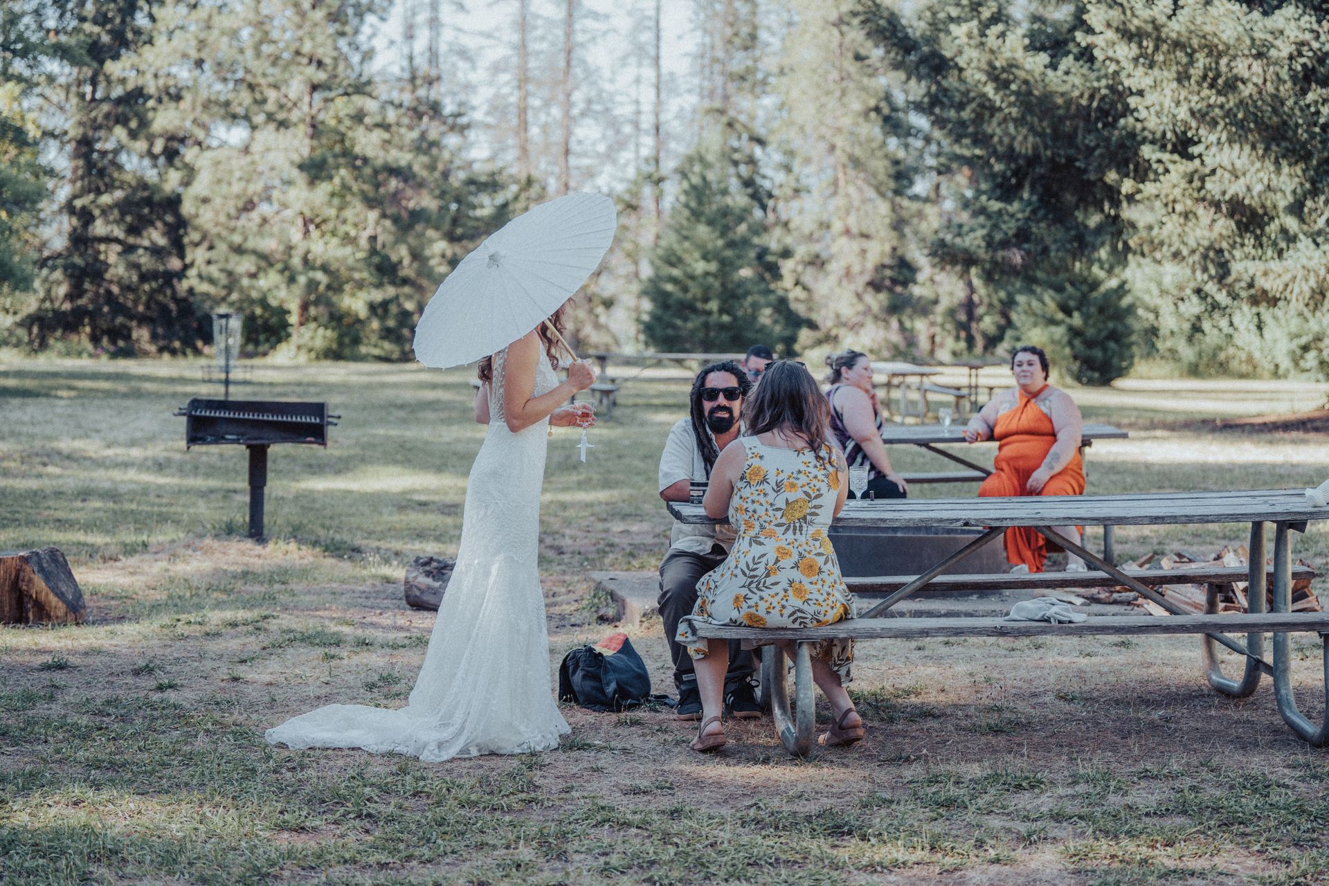 A bride is standing in front of a group of people sitting at picnic tables in a park.