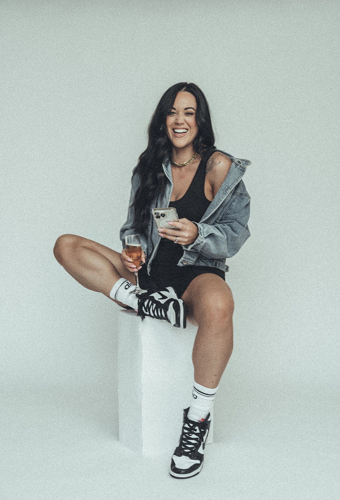 A woman is sitting on a cube holding a glass of wine and a cell phone.