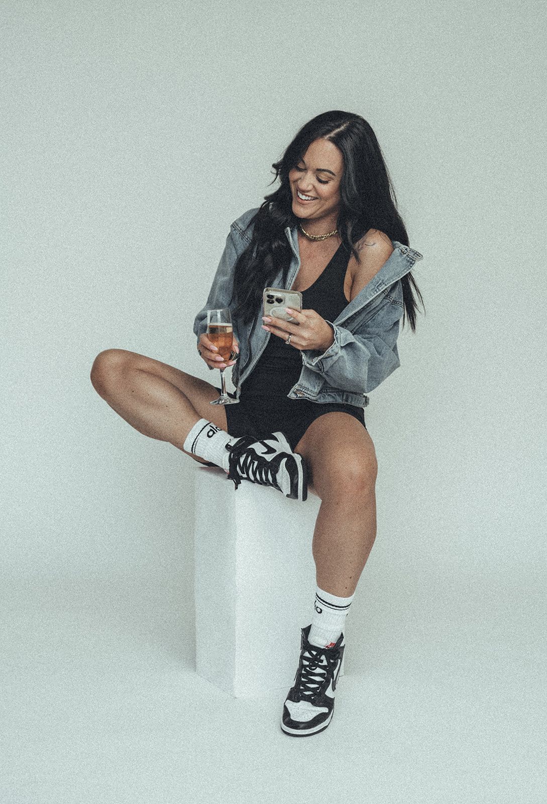 A woman is sitting on a cube holding a glass of wine and a cell phone.
