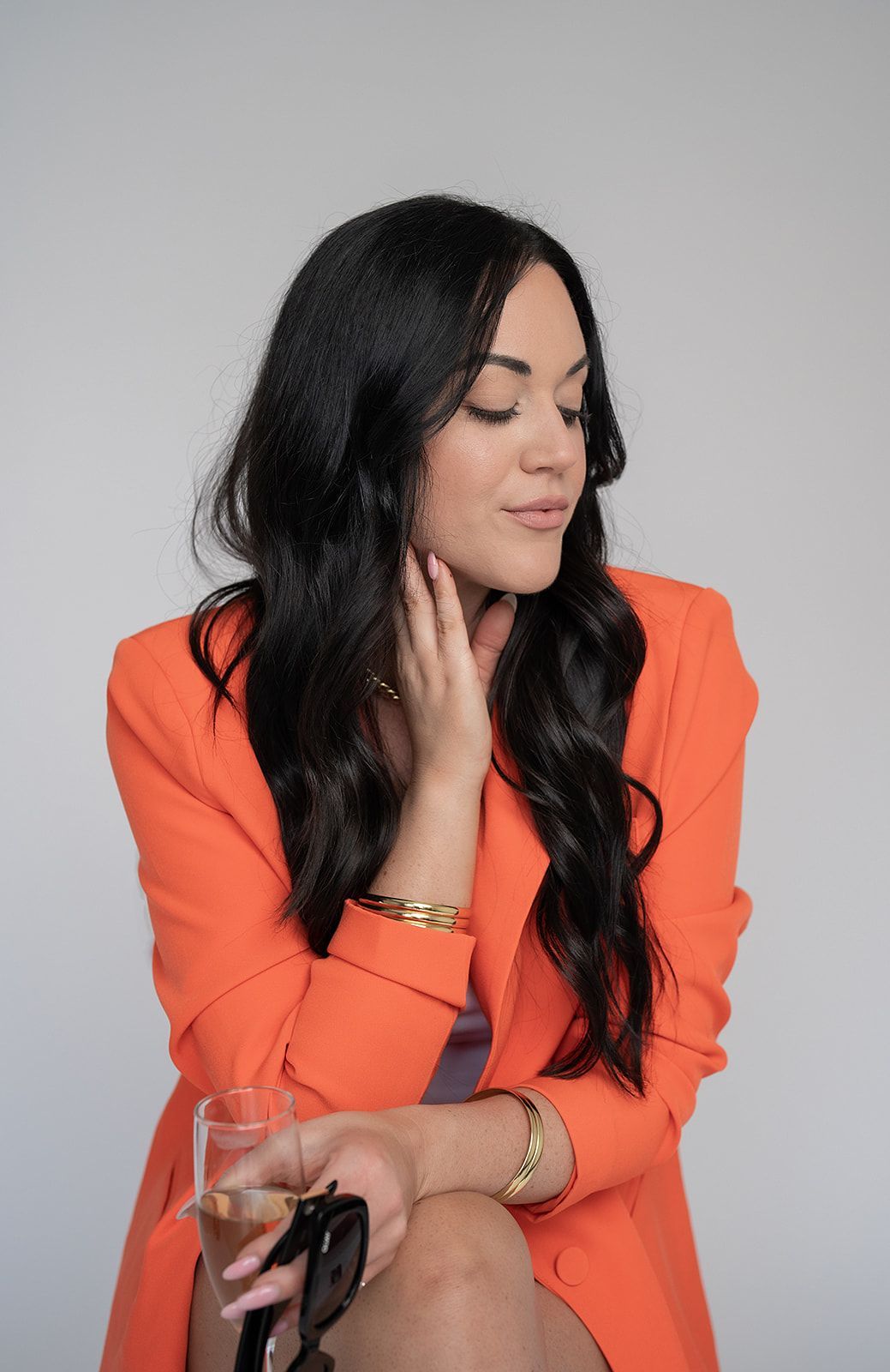 A woman in an orange jacket is holding a glass of wine and sunglasses.