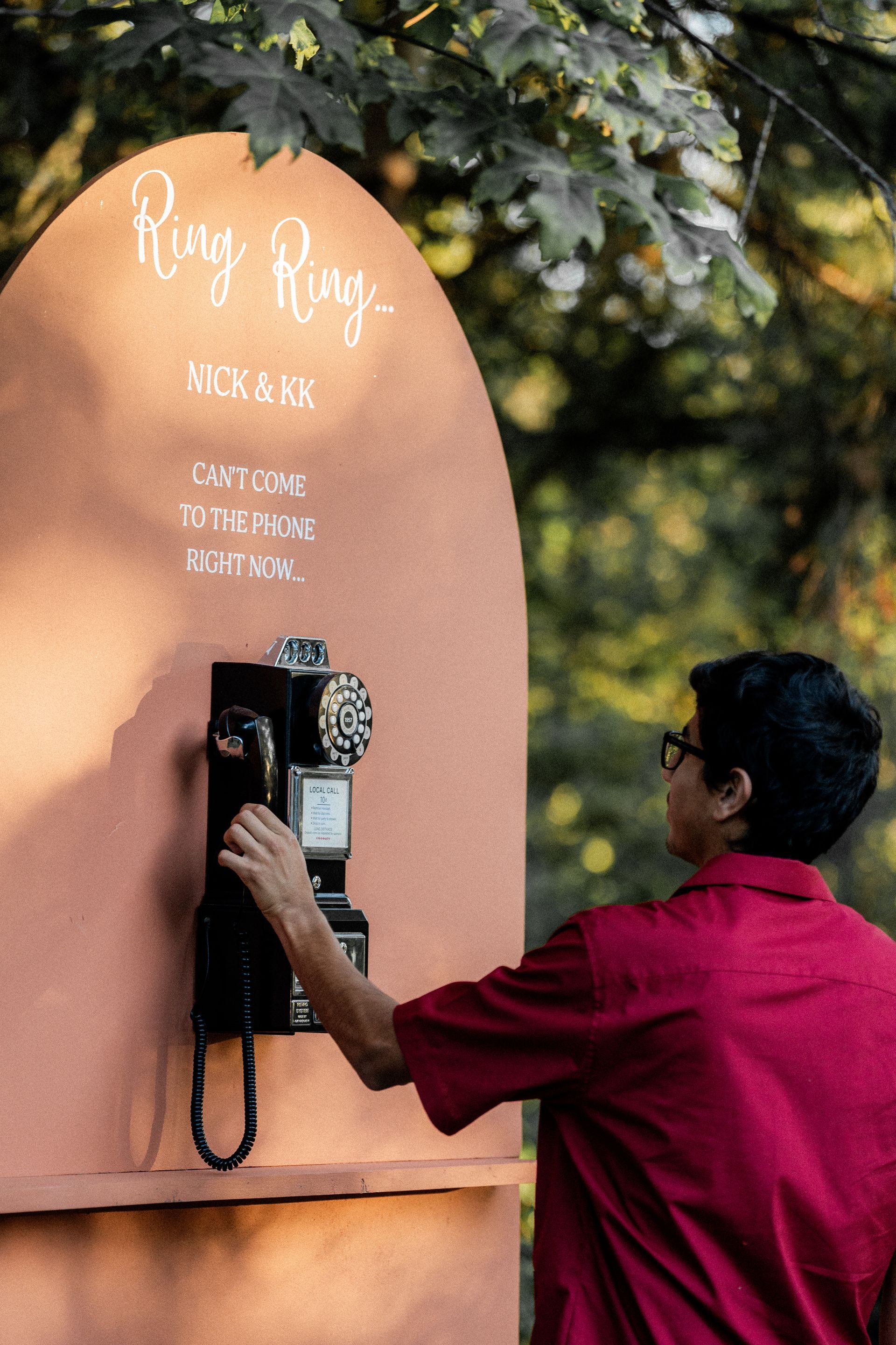 A man in a red shirt is talking on a pay phone.