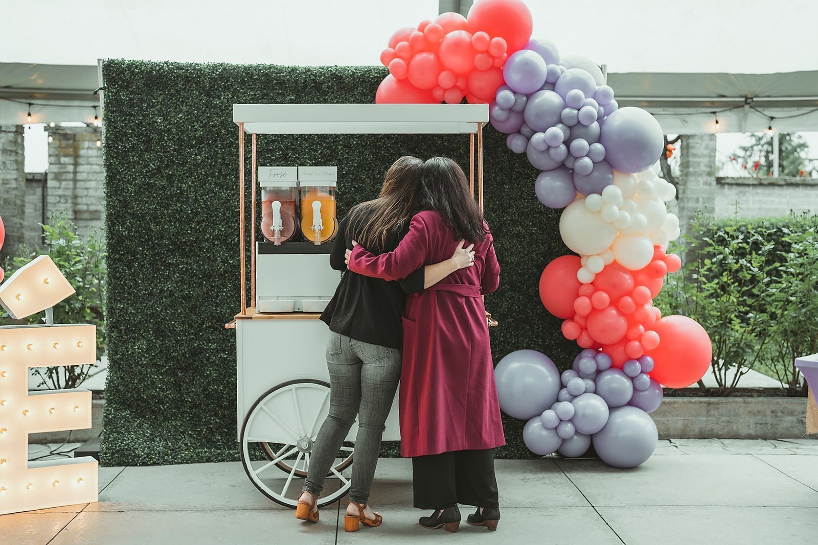 Two women are hugging in front of a cart decorated with balloons.