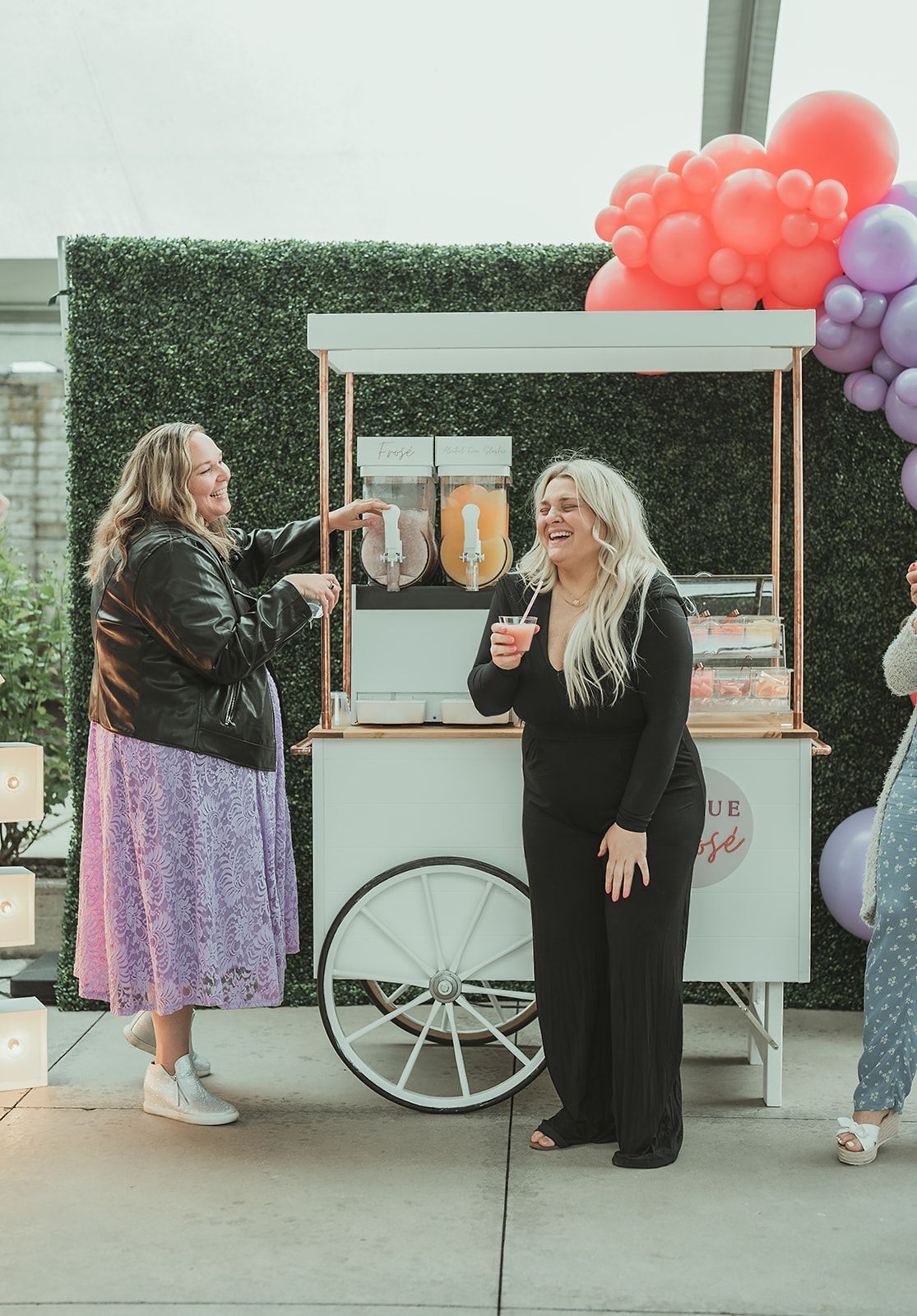 Two women are standing next to a cart with drinks and balloons.