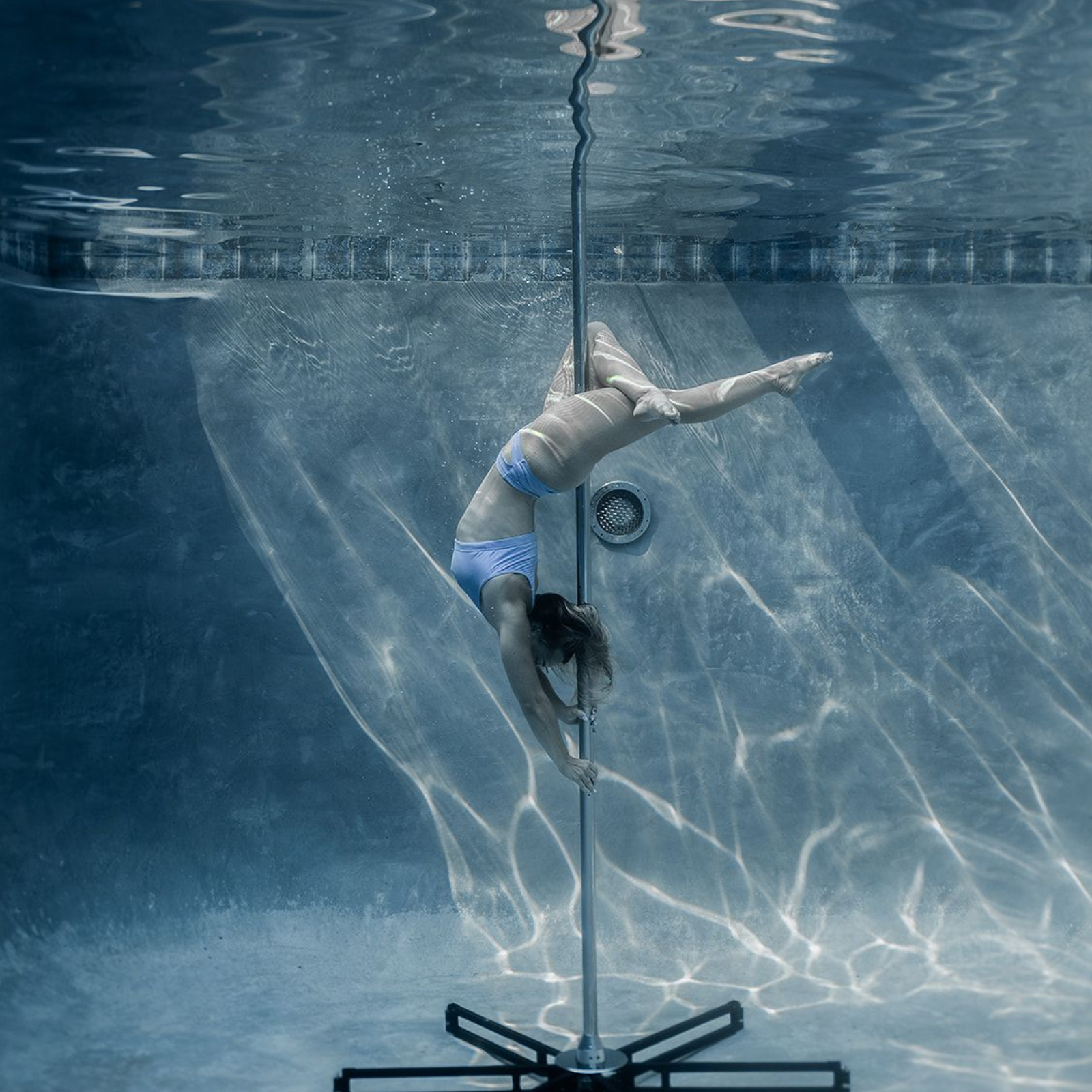 Woman underwater on a pole, wearing blue bikini, performing a pose in a pool.