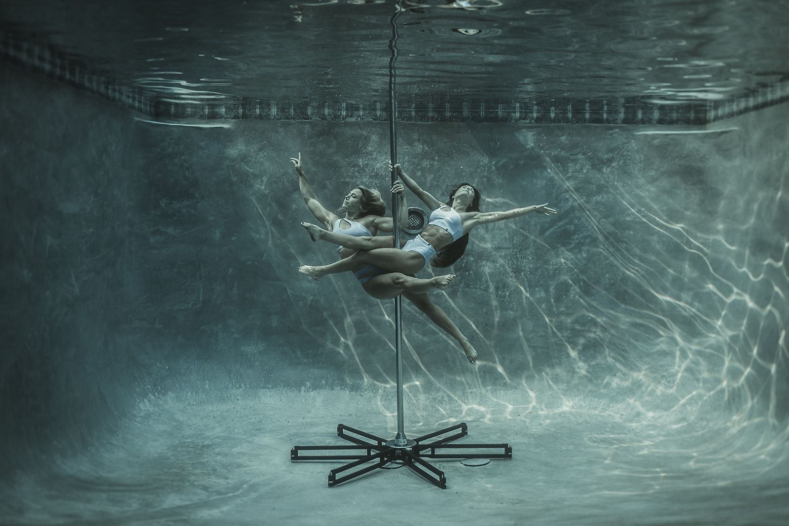 Two women are doing aerial acrobatics underwater in a pool.