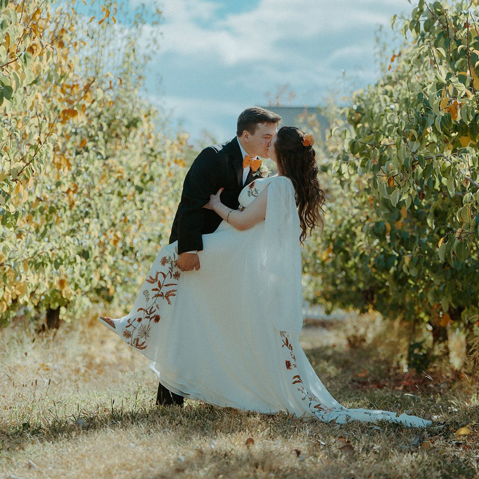 Groom dips bride for a kiss in a sunny orchard, fall leaves in the background. She wears a white dress with red floral embroidery.