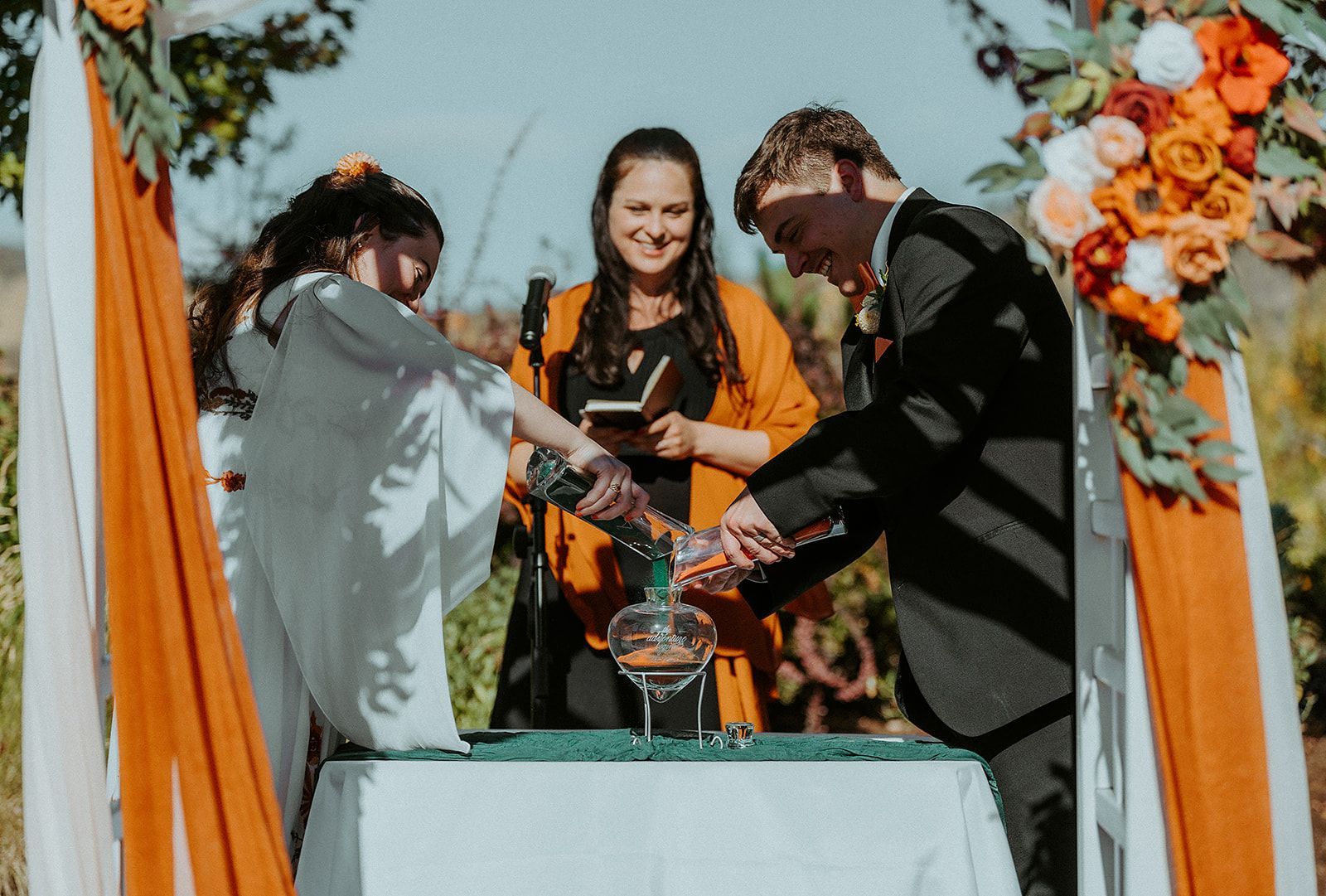 A bride and groom are pouring liquid into a glass during their wedding ceremony.