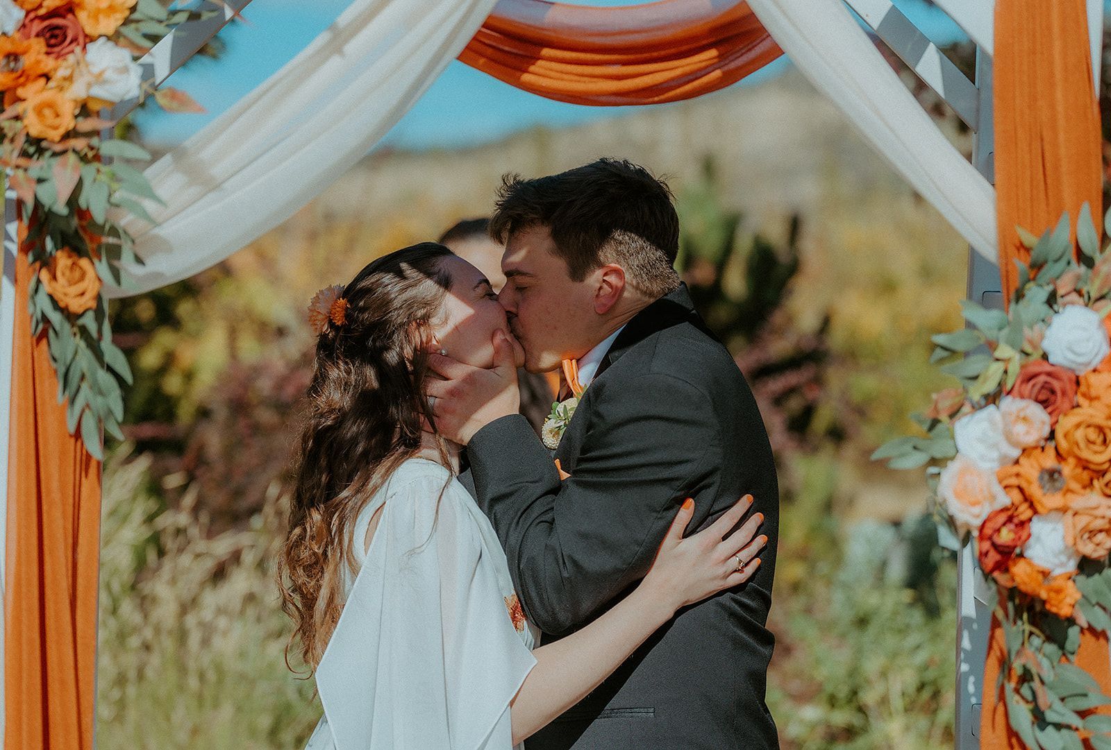 A bride and groom are kissing under an arch at their wedding.