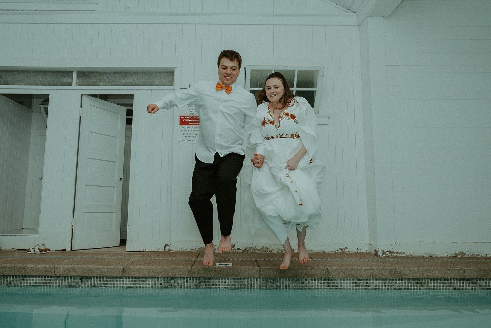 A bride and groom are jumping into a swimming pool.