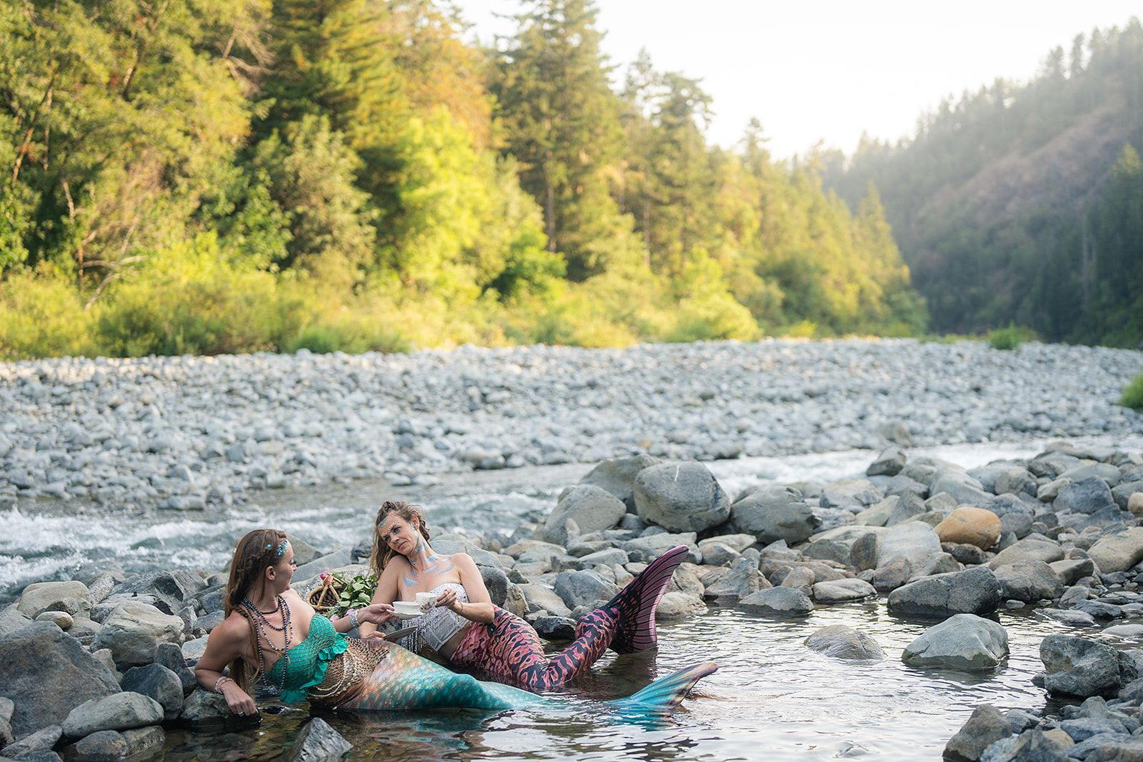 Two mermaids are laying on the rocks in a river.