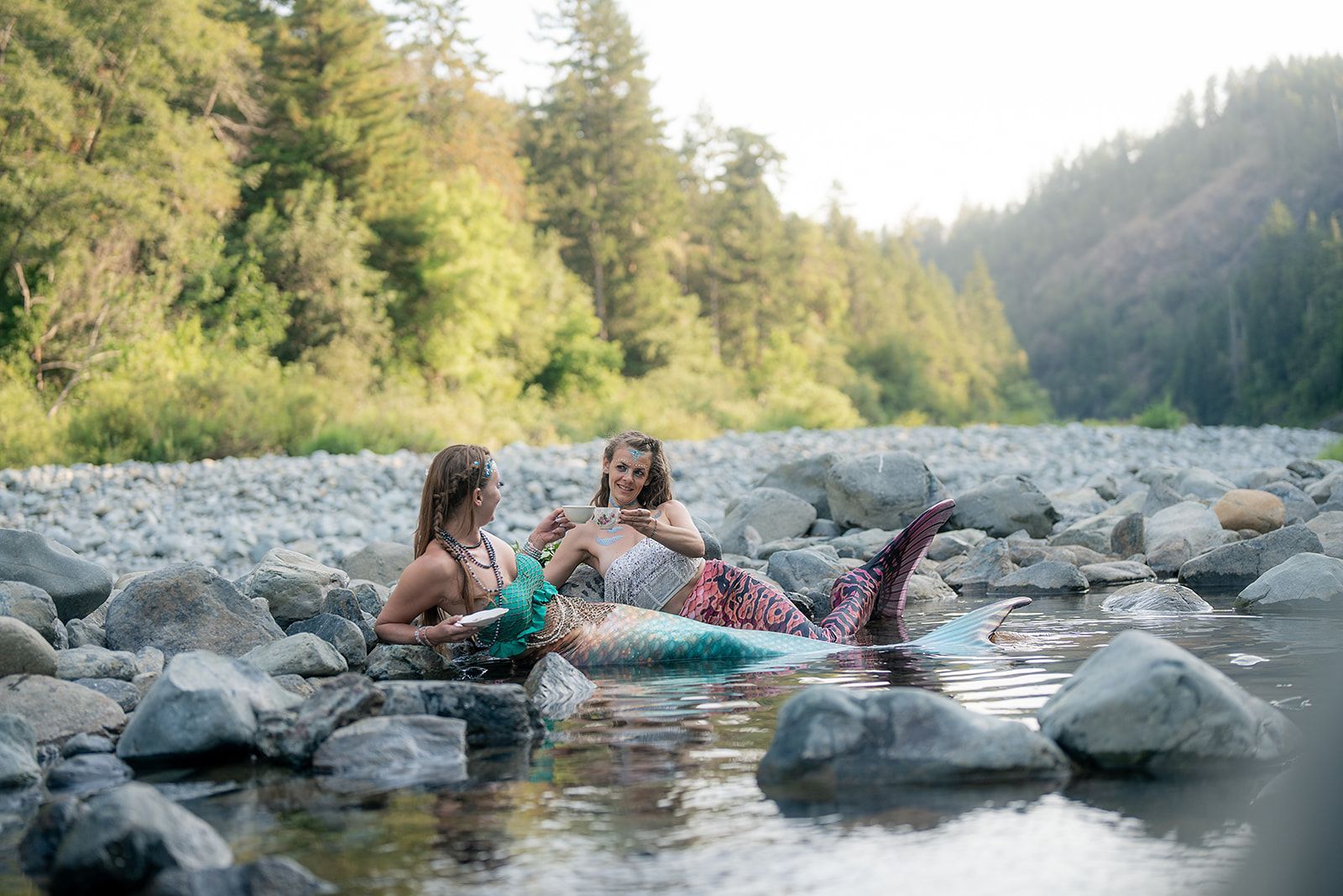 Two mermaids are sitting on rocks in a river.