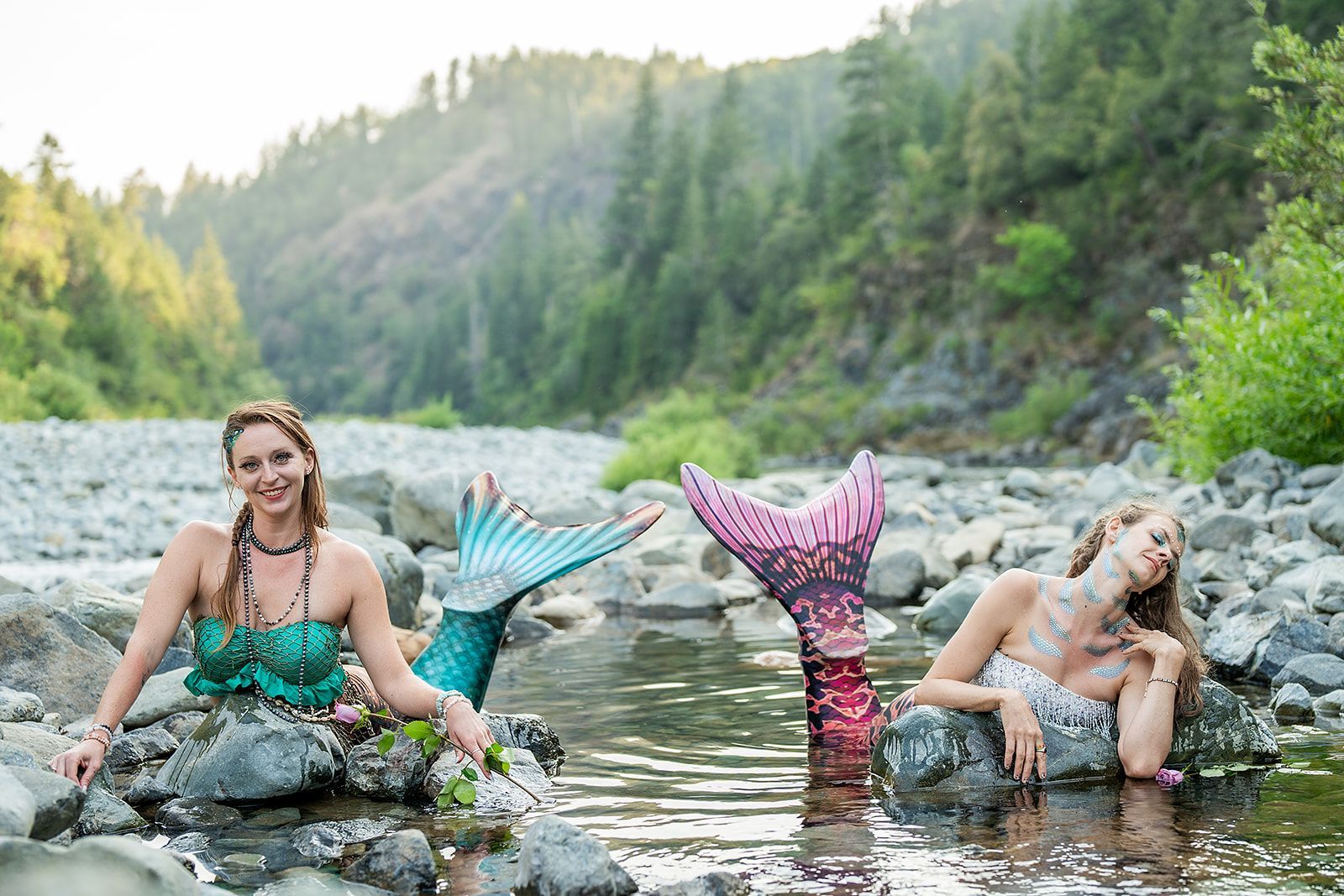 Two mermaids are sitting on rocks in a river.