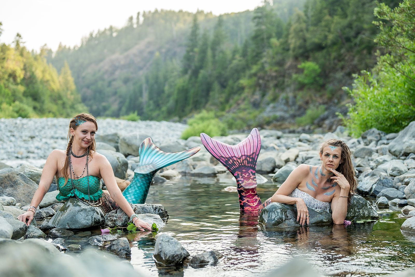 Two mermaids are sitting on rocks in a river.