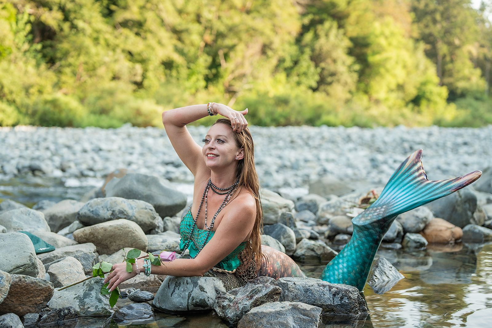 A woman dressed as a mermaid is sitting on a rock in a river.