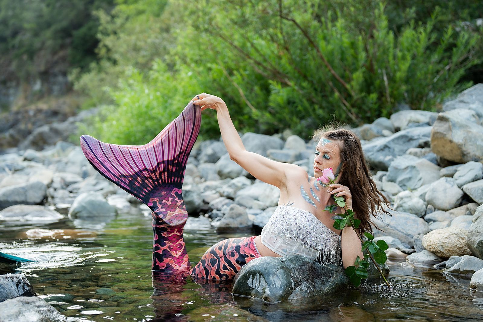 A mermaid is sitting on a rock in a river holding a flower.