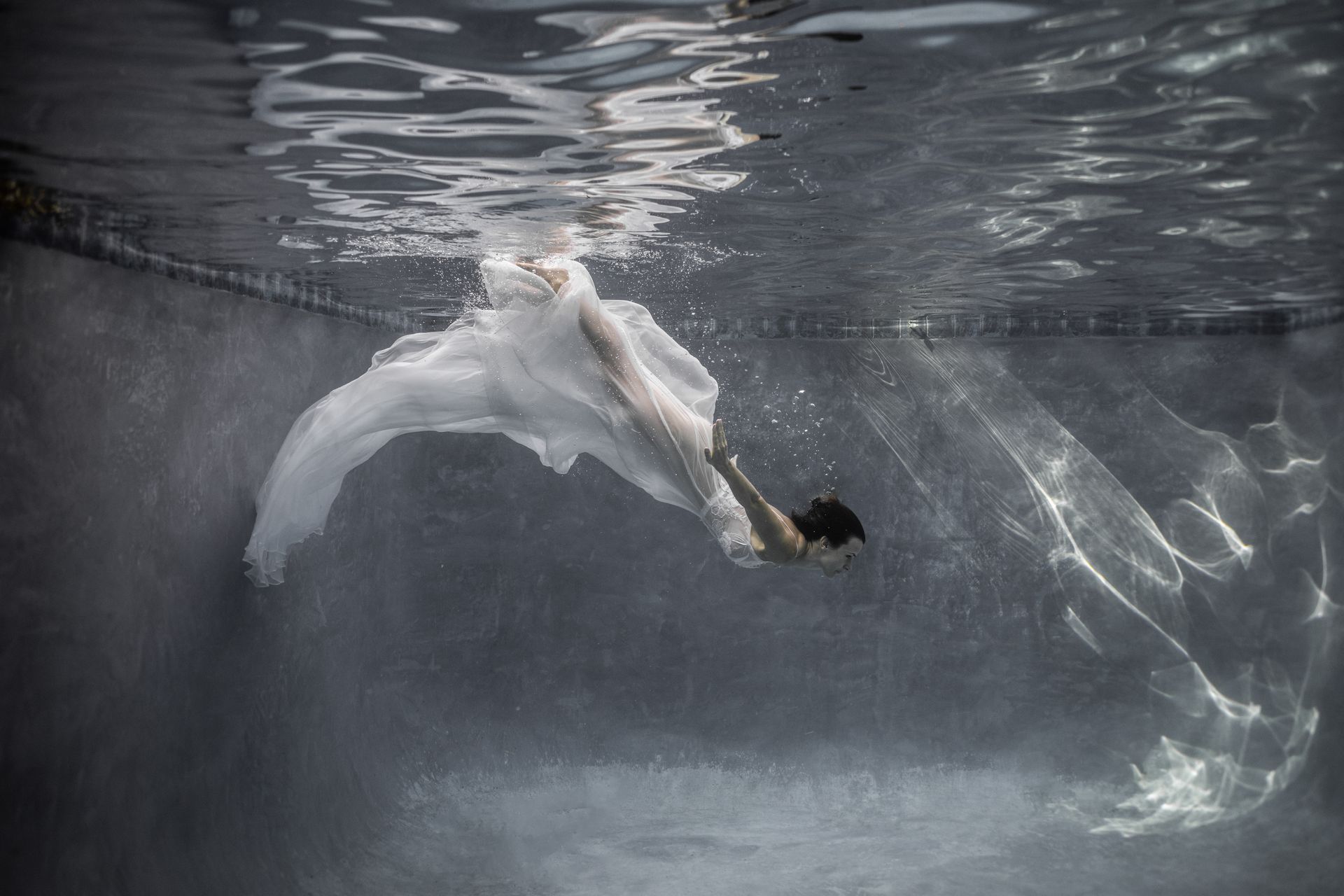 A woman in a white dress is swimming underwater in a pool.