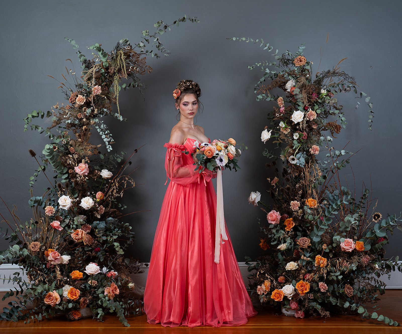 A woman in a red dress is standing in front of a floral arch holding a bouquet of flowers.