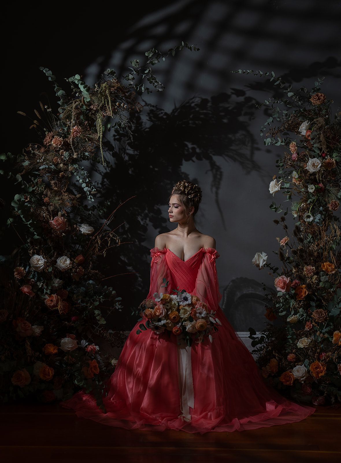A woman in a red dress is sitting in front of a wall of flowers.