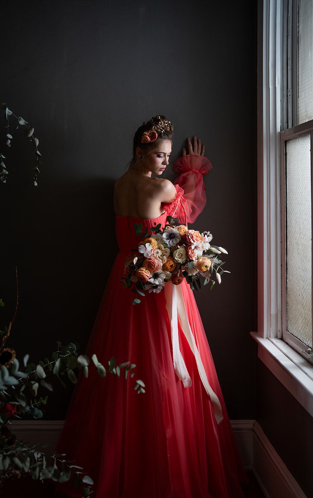 A woman in a red dress is standing in front of a window holding a bouquet of flowers.