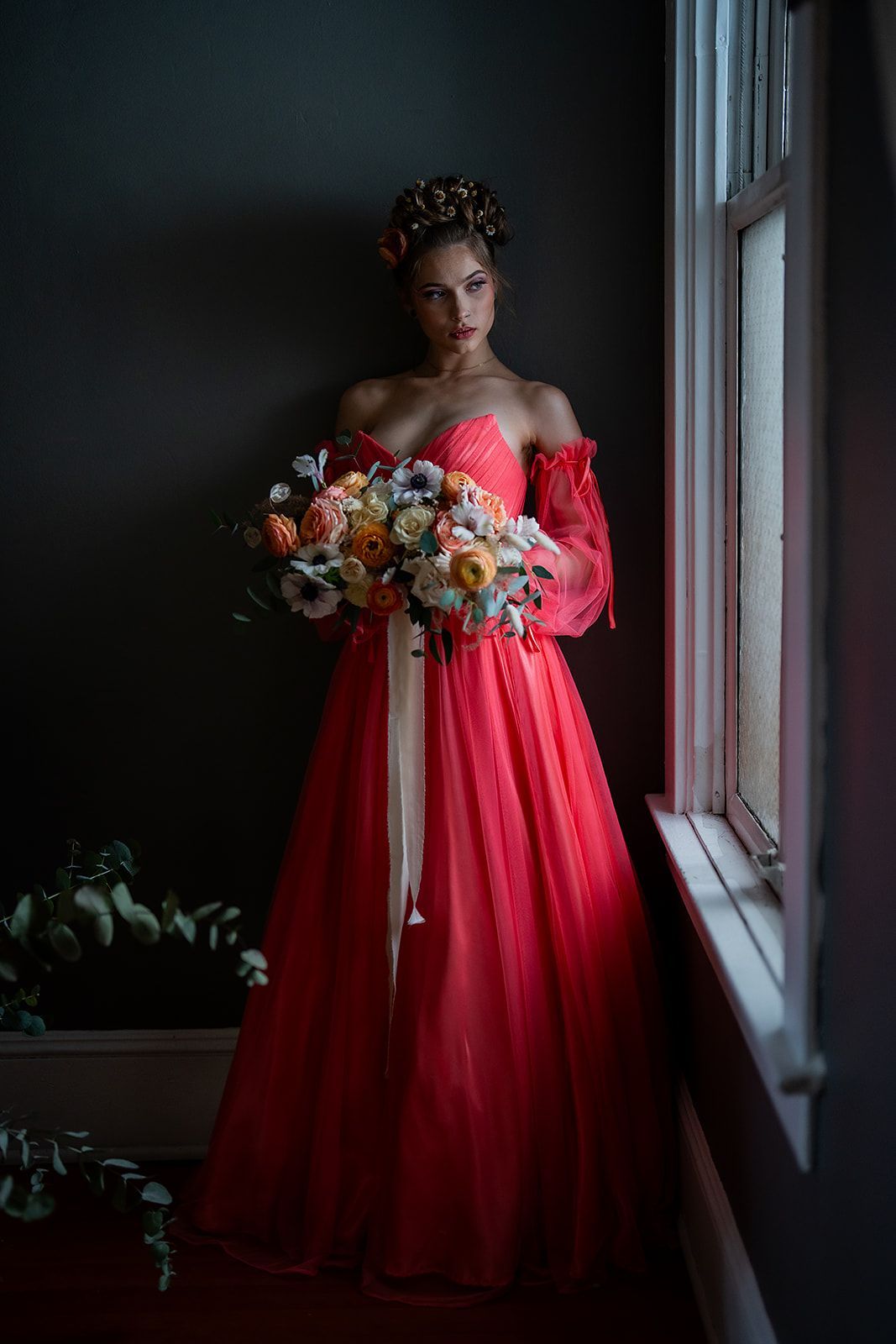 A woman in a red dress is standing in front of a window holding a bouquet of flowers.